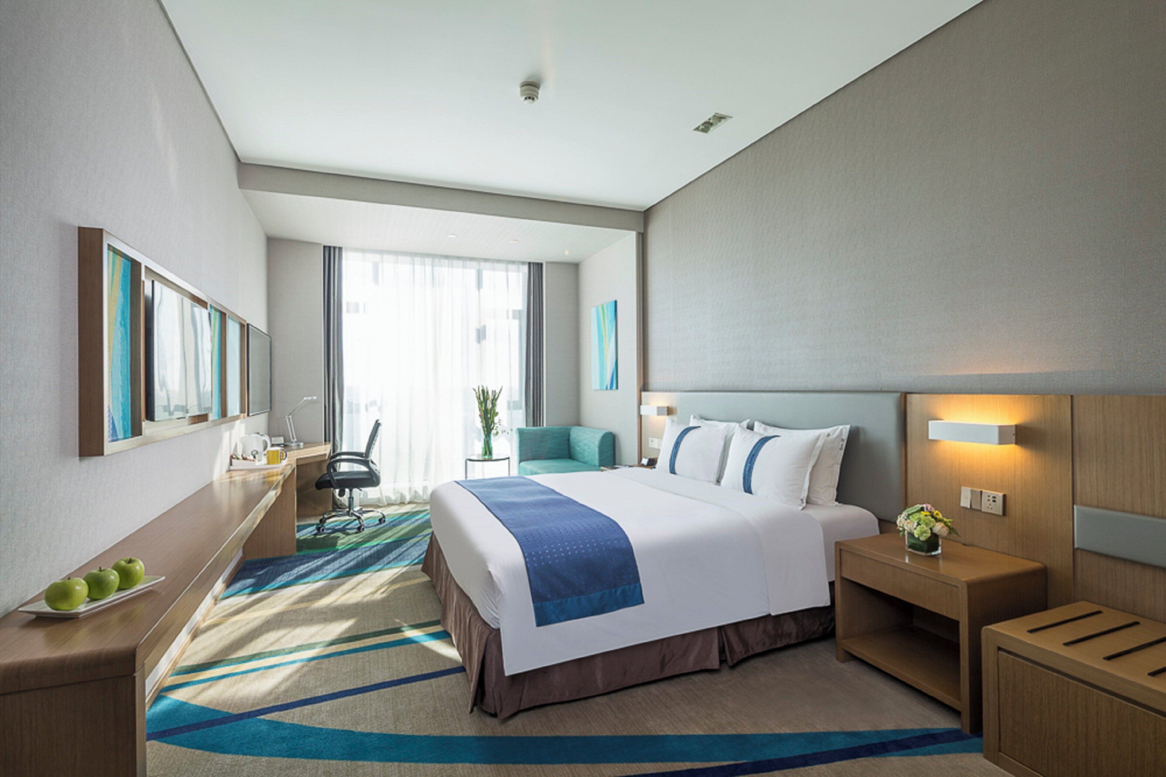 Quarto de hotel com uma cama grande, uma escrivaninha, uma cadeira, uma mesinha de cabeceira e um criado-mudo.