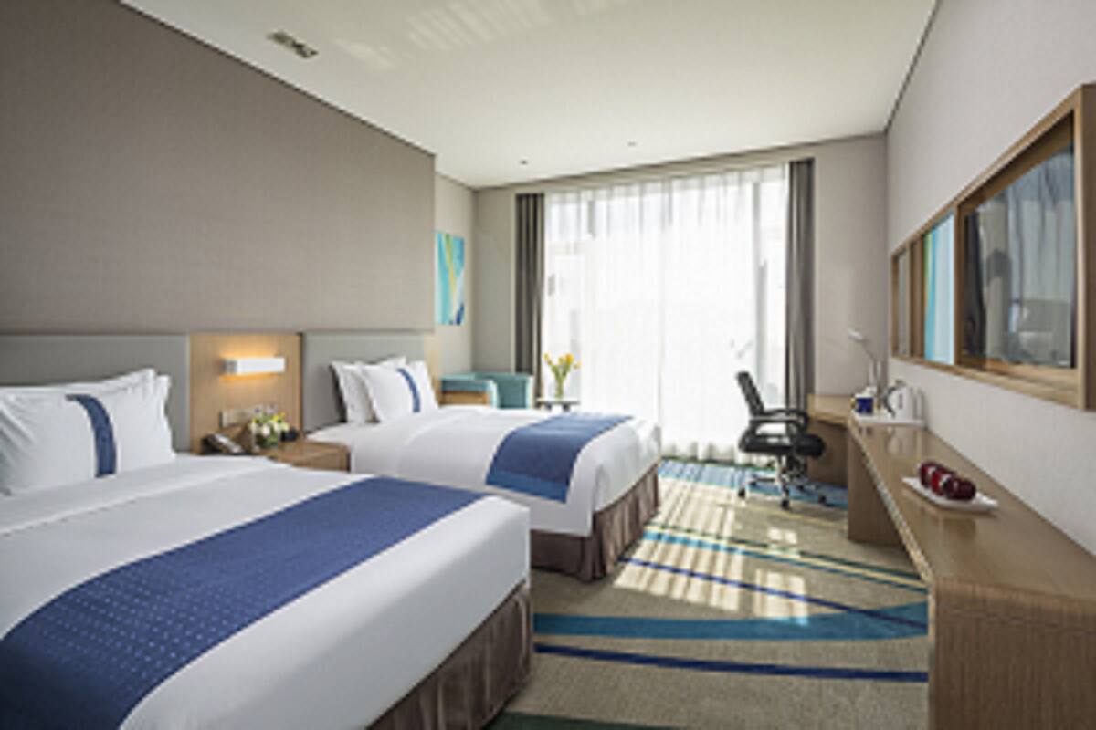 Une chambre d’hôtel avec deux lits, un bureau, une chaise, une grande fenêtre avec des rideaux et un miroir au mur.