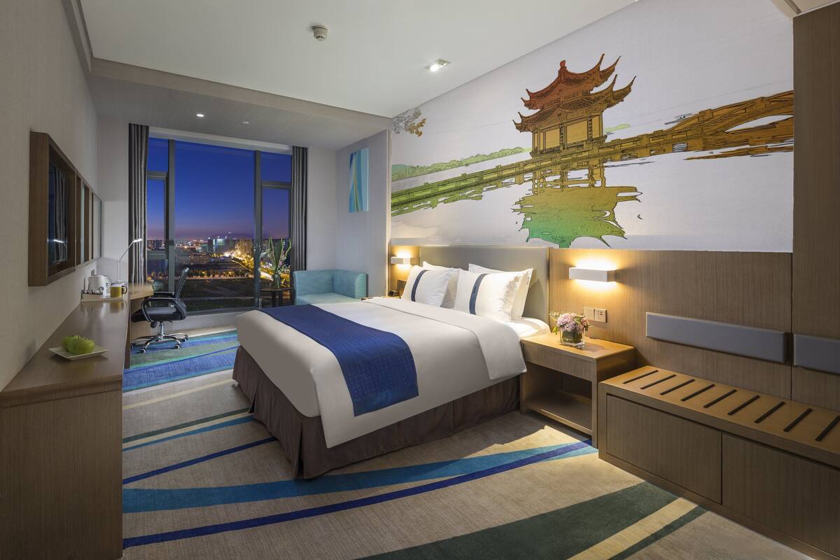 Une chambre d’hôtel moderne avec un grand lit, un bureau, une chaise et une vue sur la ville la nuit.