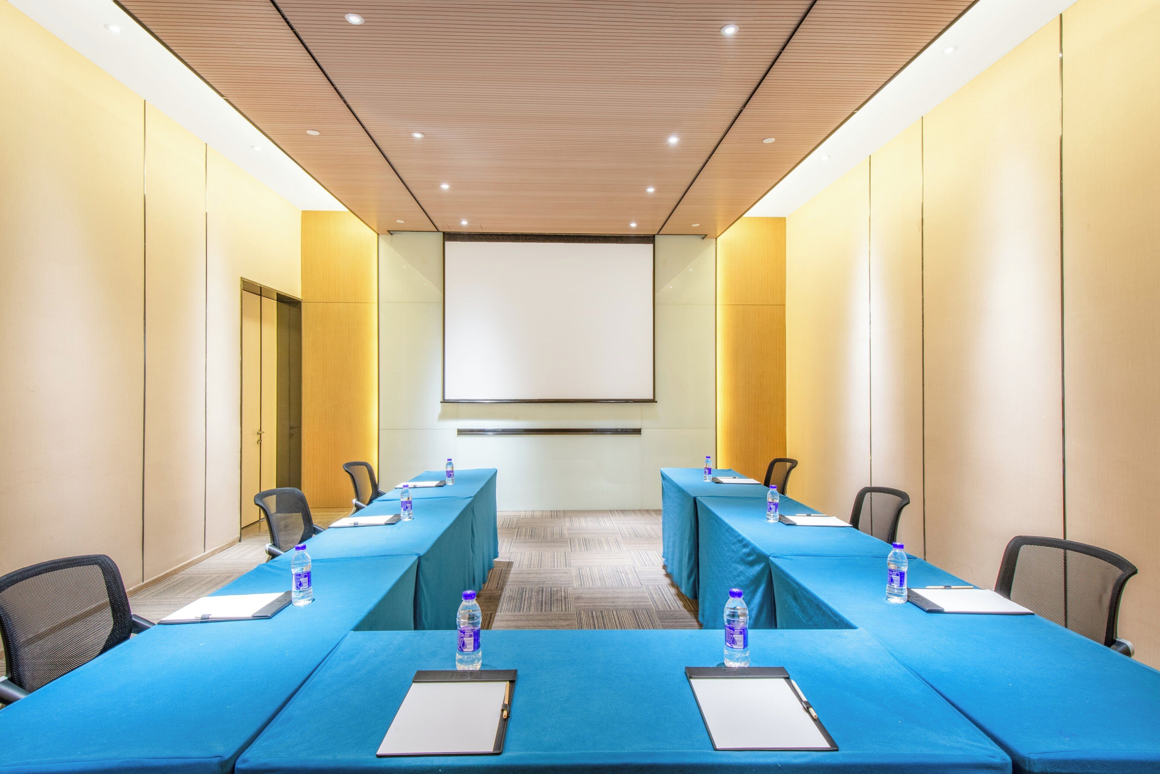 Un salón de conferencias con una mesa larga, sillas, una pantalla de proyector y botellas de agua.