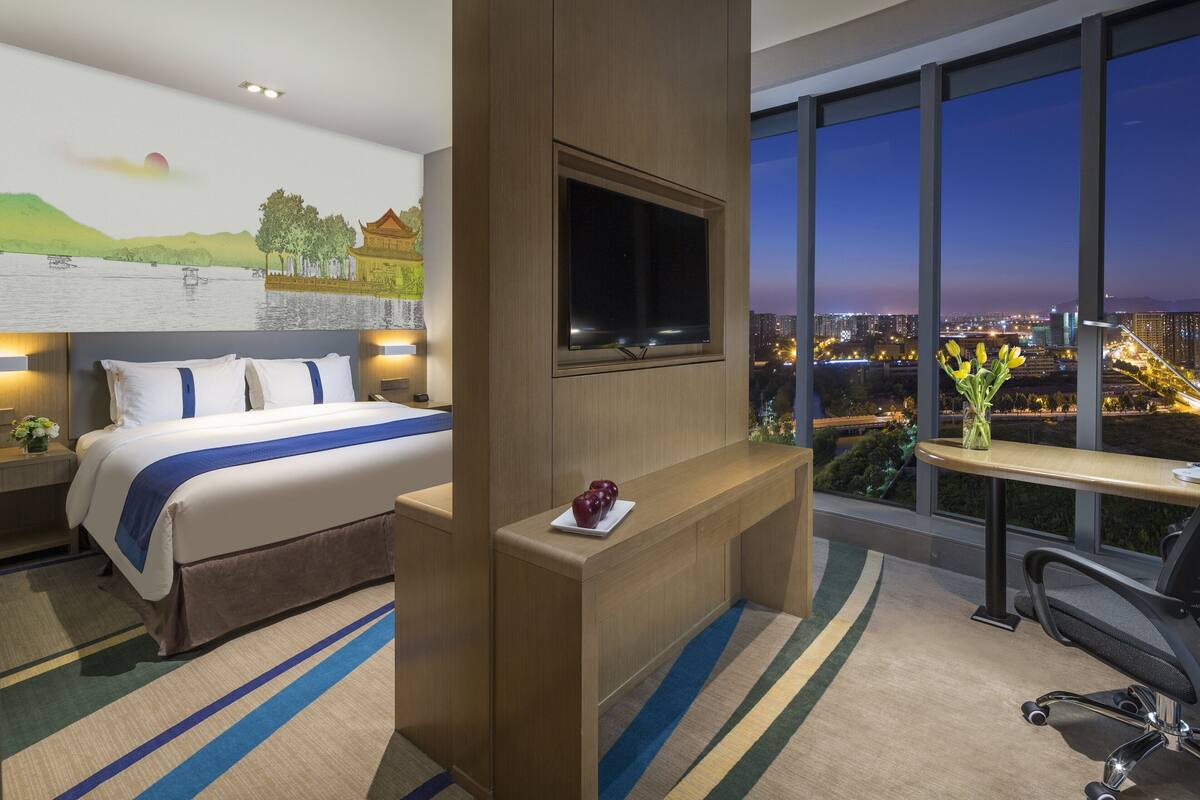 Une chambre d’hôtel avec un grand lit, un bureau, un téléviseur fixé au mur et une vue sur la ville au crépuscule.