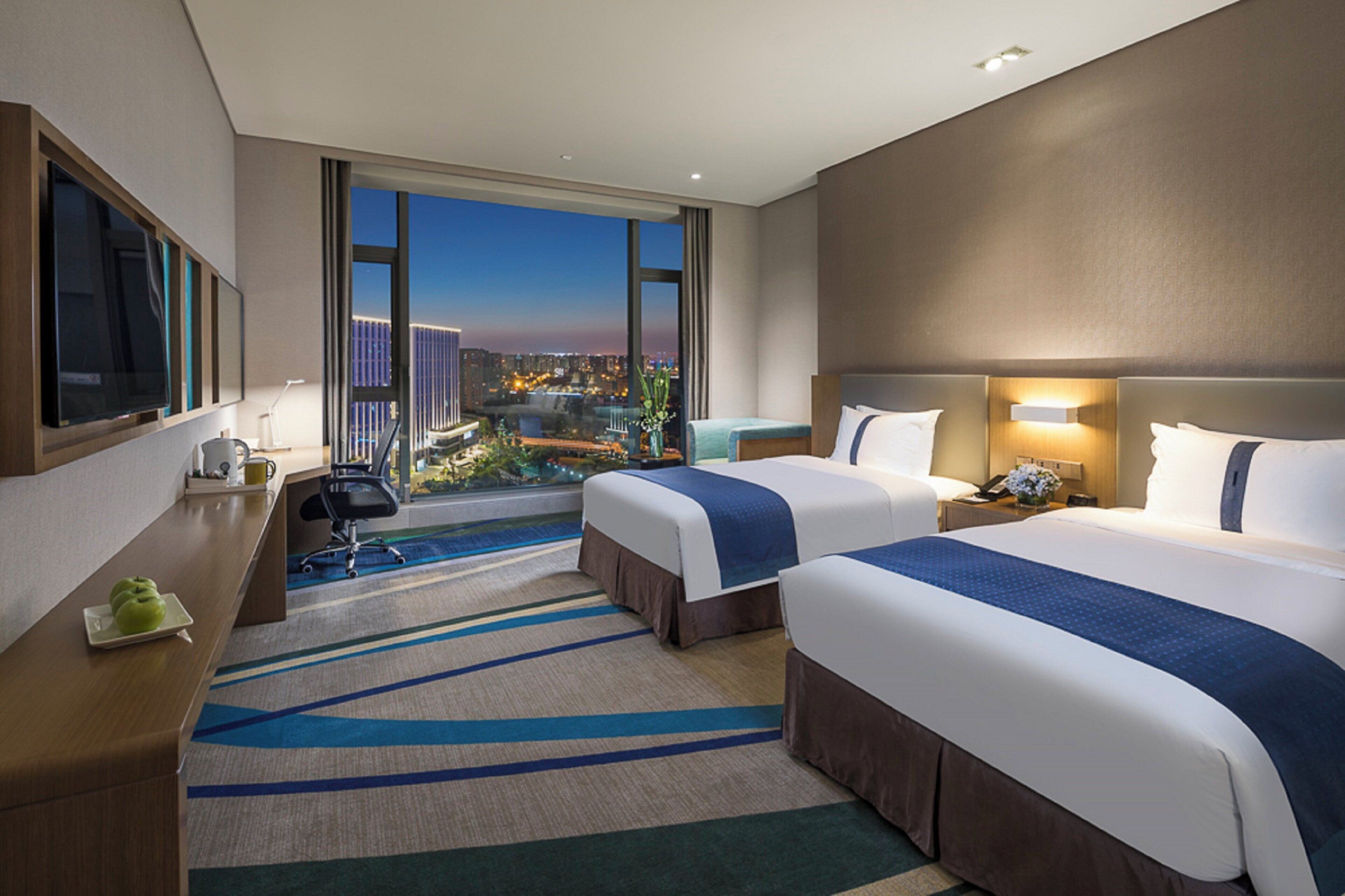 A hotel room with two beds, a desk, a chair, a TV, and a view of the city at dusk.