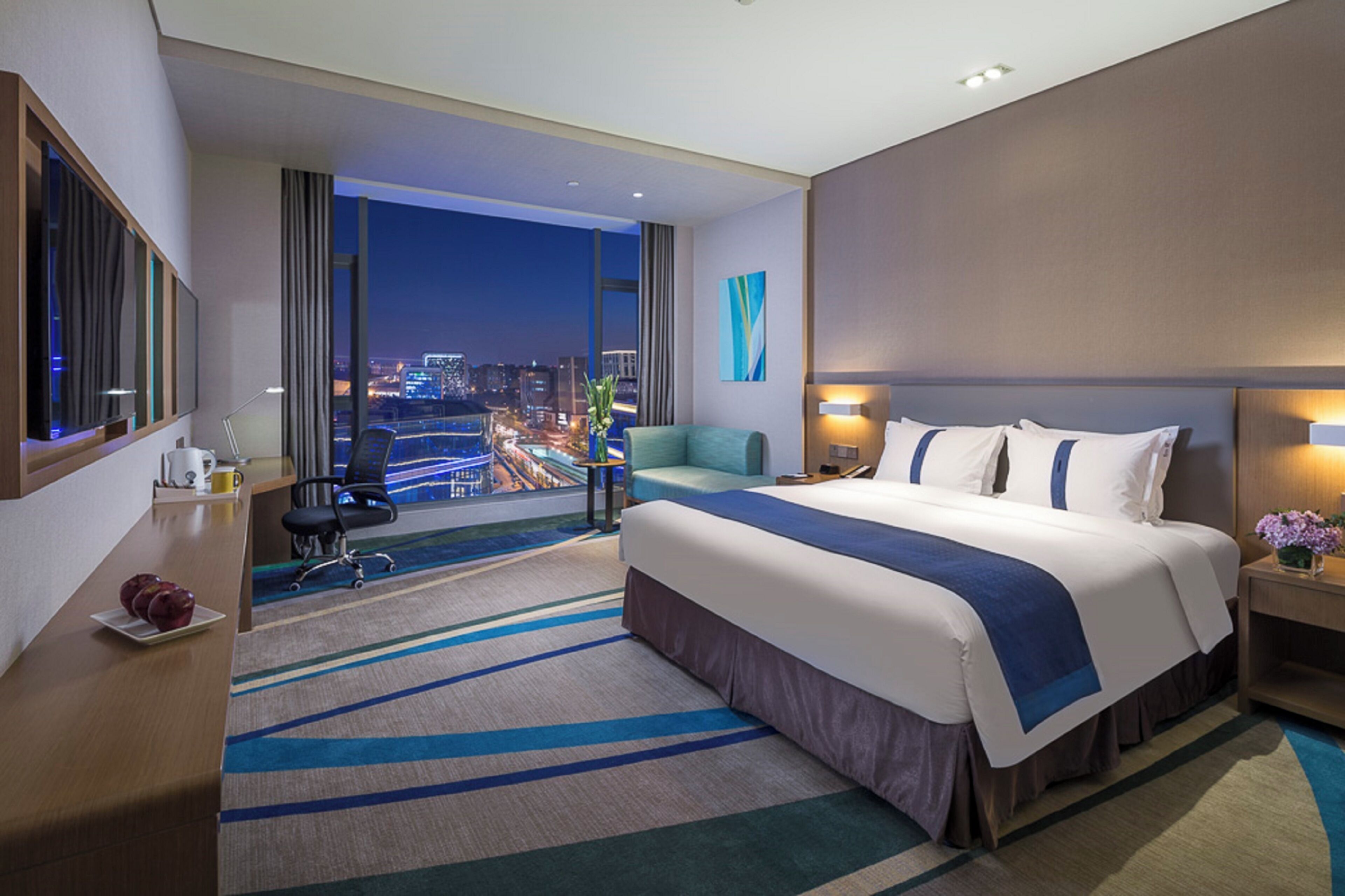 A hotel room with a large bed, a desk, a chair, a view of the city at night, and a window with curtains.
