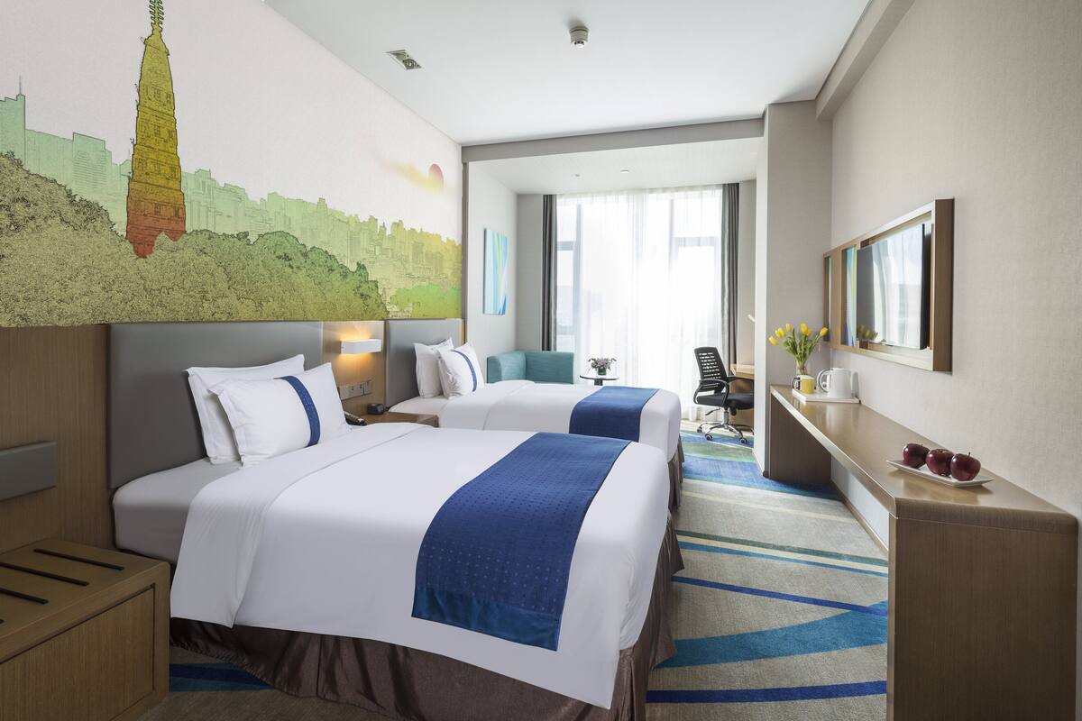 Une chambre d’hôtel avec deux lits, un bureau, une chaise, une grande fenêtre et une fresque représentant un paysage urbain sur le mur.