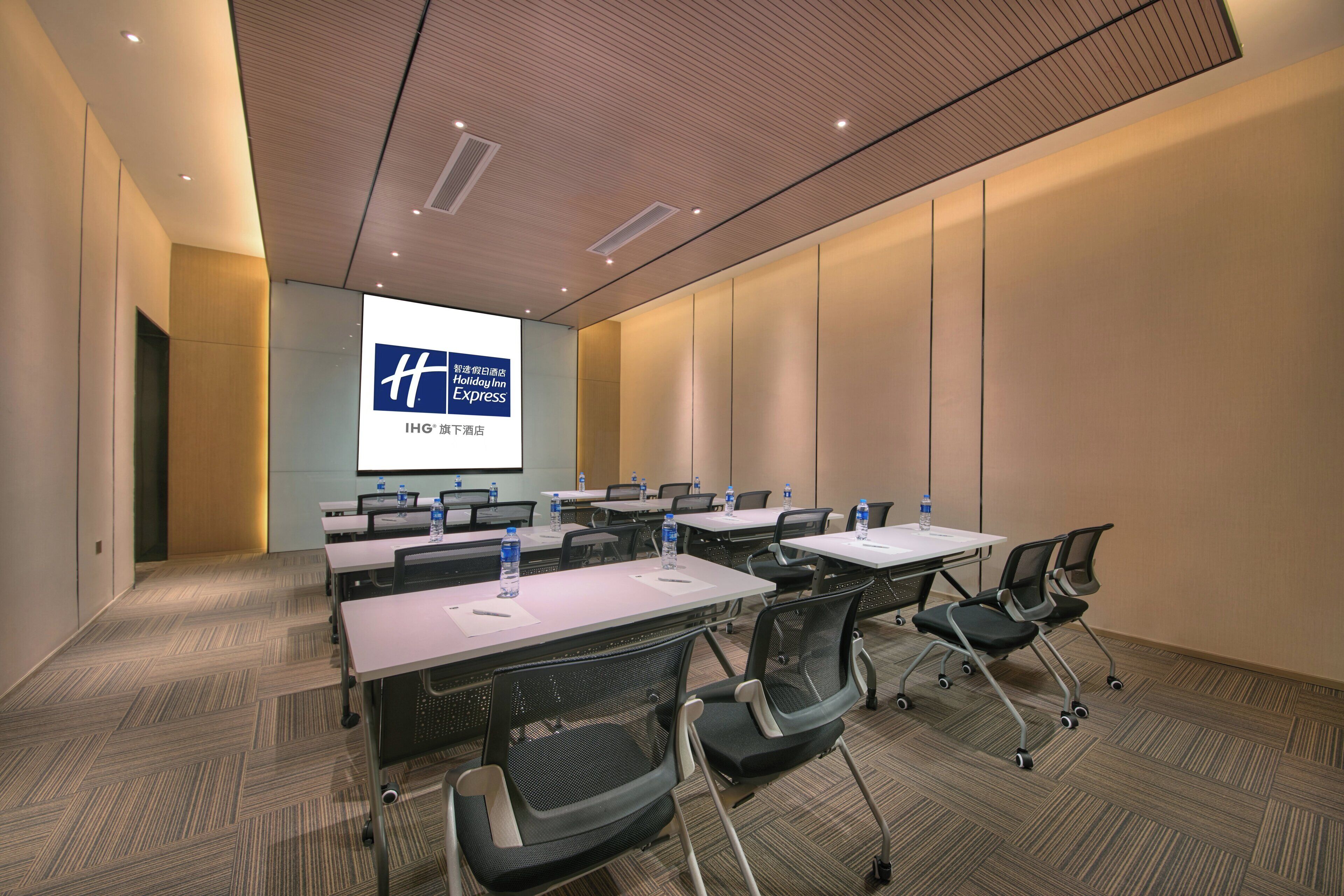 A conference room with a large screen displaying a hotel logo, multiple tables with chairs, and a carpeted floor.