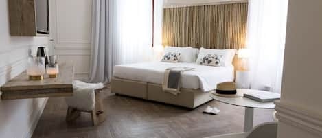 A modern hotel room with a large bed, a wooden headboard, a small desk with a lamp, a chair, and a herringbone-patterned floor.
