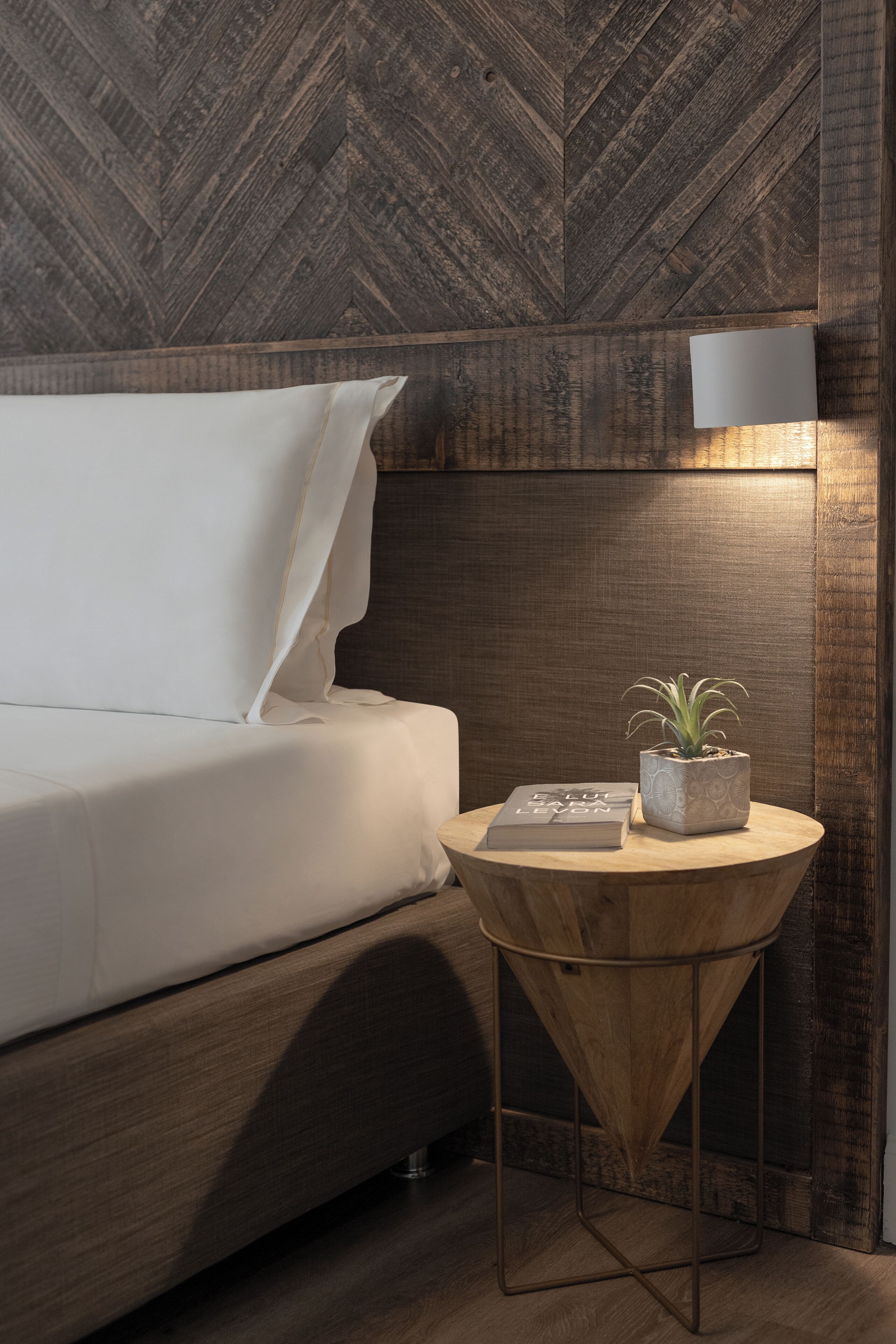 A neatly made bed with white linens, a small round wooden side table with a potted plant, and a wall-mounted light fixture.
