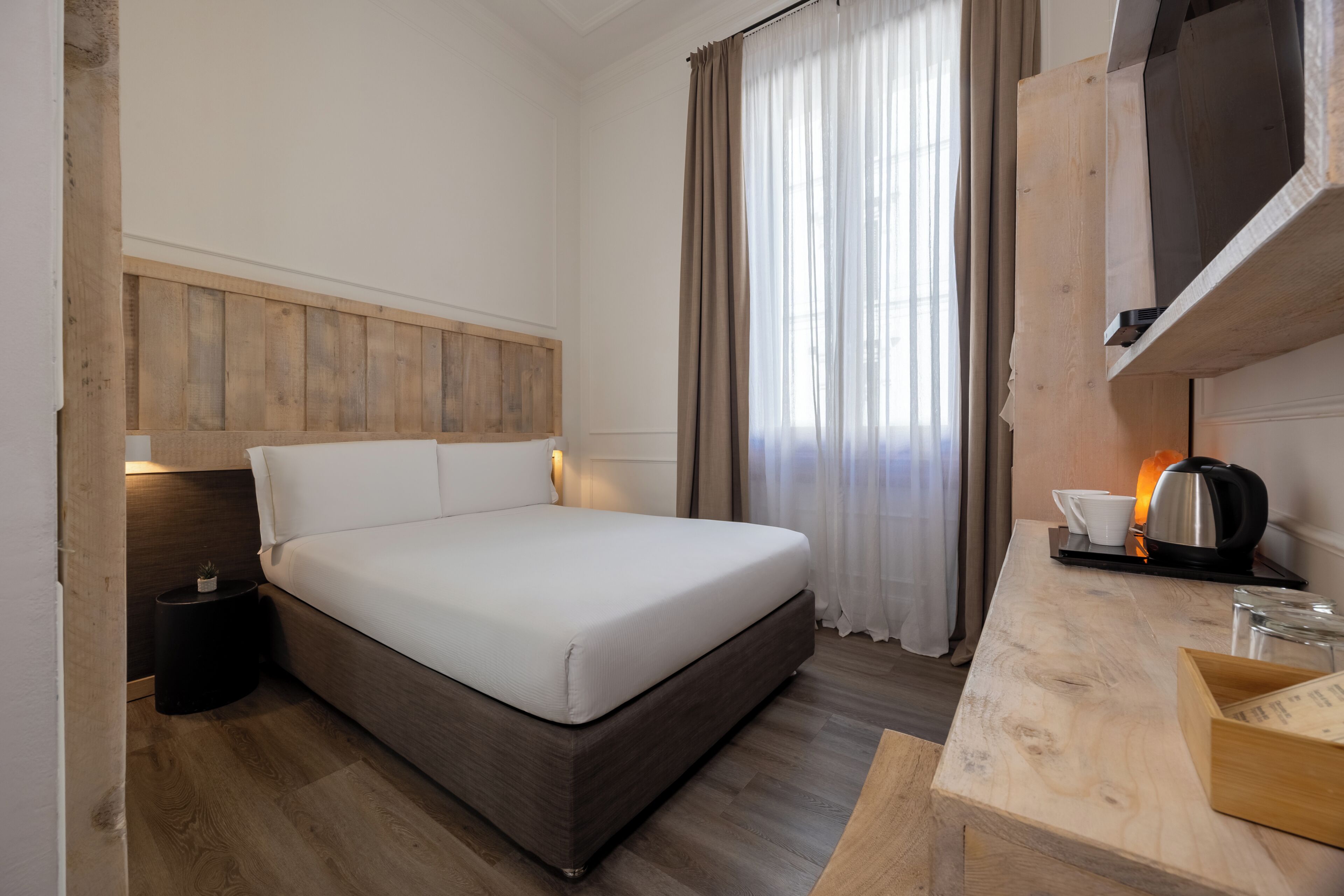 A hotel room with a bed, bedside table, lamp, curtains, and a wooden desk with a kettle.