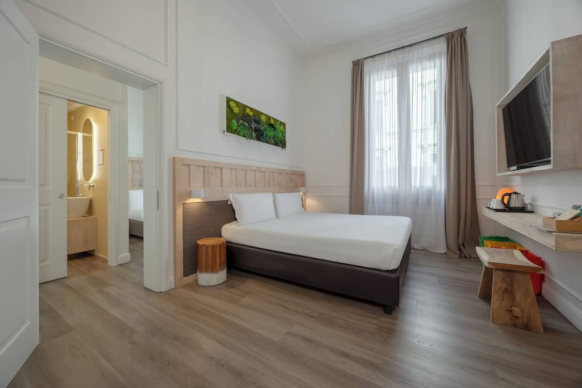 A hotel room with a bed, a TV, a painting, a window with curtains, and a wooden floor.