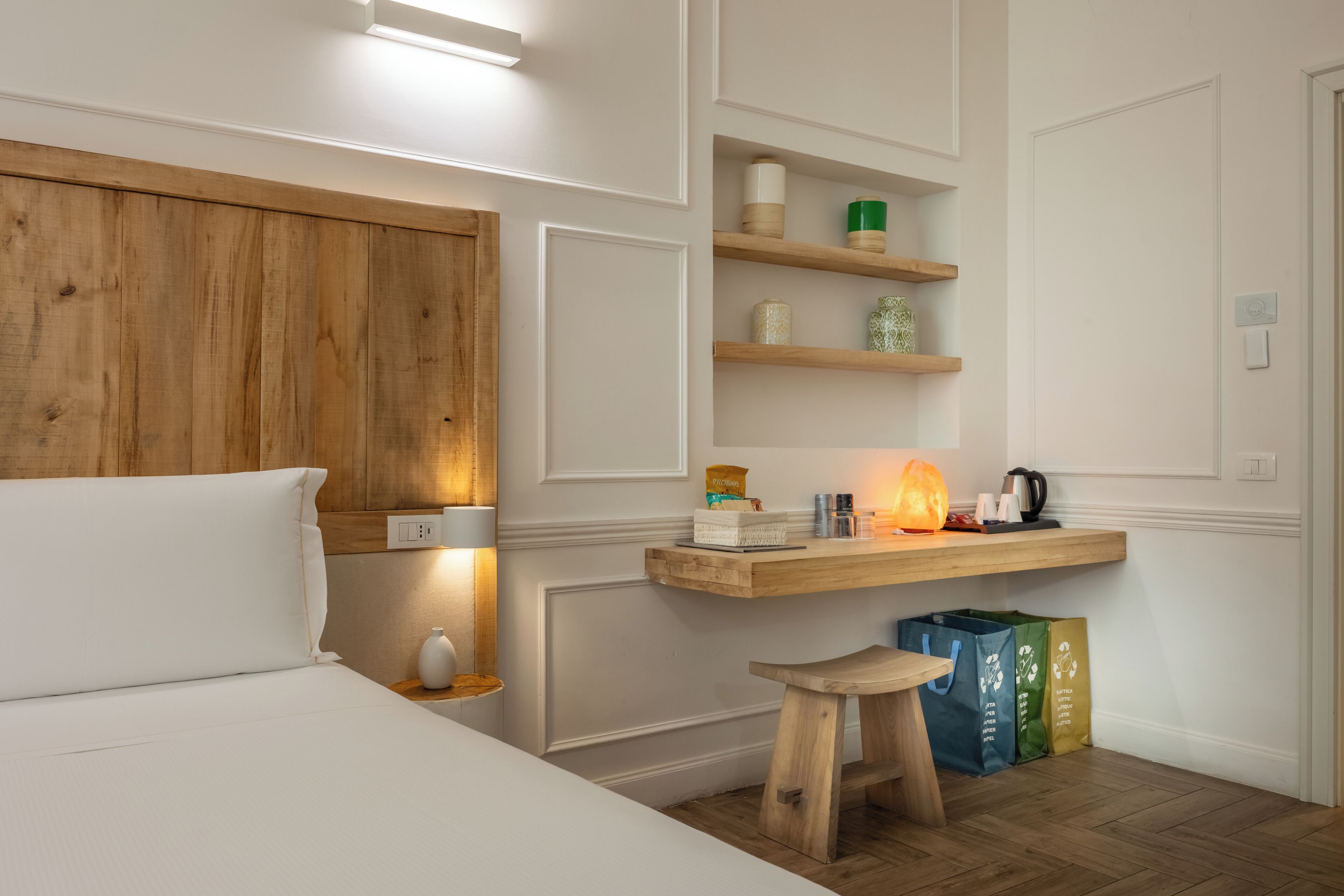 A room with a bed, a wooden headboard, a nightstand with a lamp, a wooden stool, and a shelf with various items.
