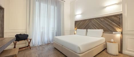 A hotel room with a large bed, a wooden desk, a chair, a small table, a lamp, and a window with sheer curtains.