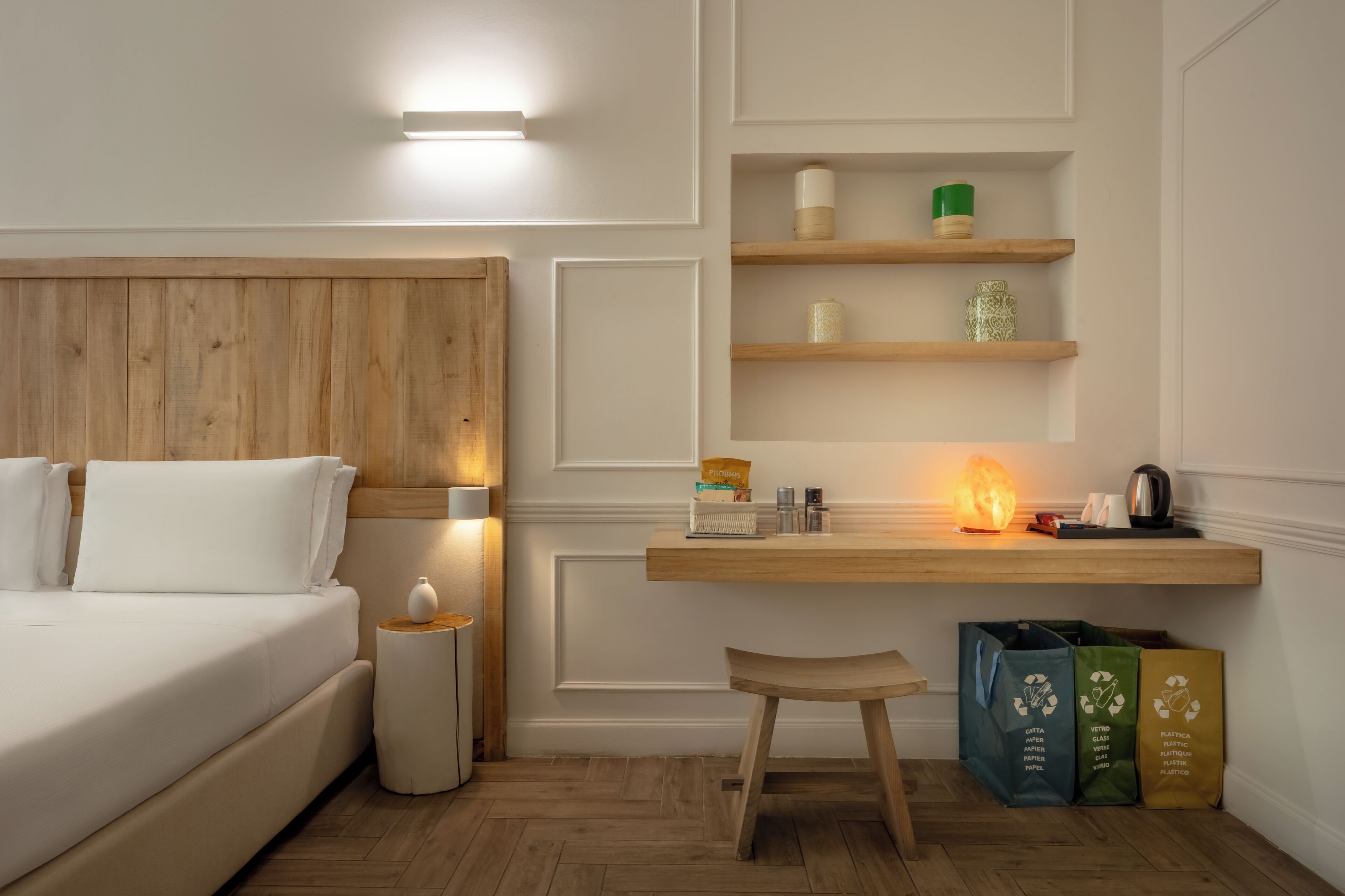 A hotel room with a wooden headboard, a bedside table with a lamp, a desk with a shelf, and a chair.