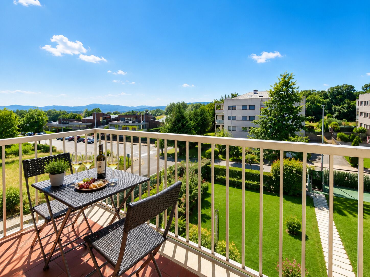 A balcony with a table, chairs, and a bottle of wine, overlooking a well-maintained lawn and buildings.