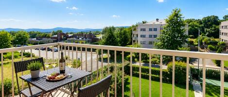 A balcony with a table, chairs, and a bottle of wine, overlooking a well-maintained lawn and buildings.