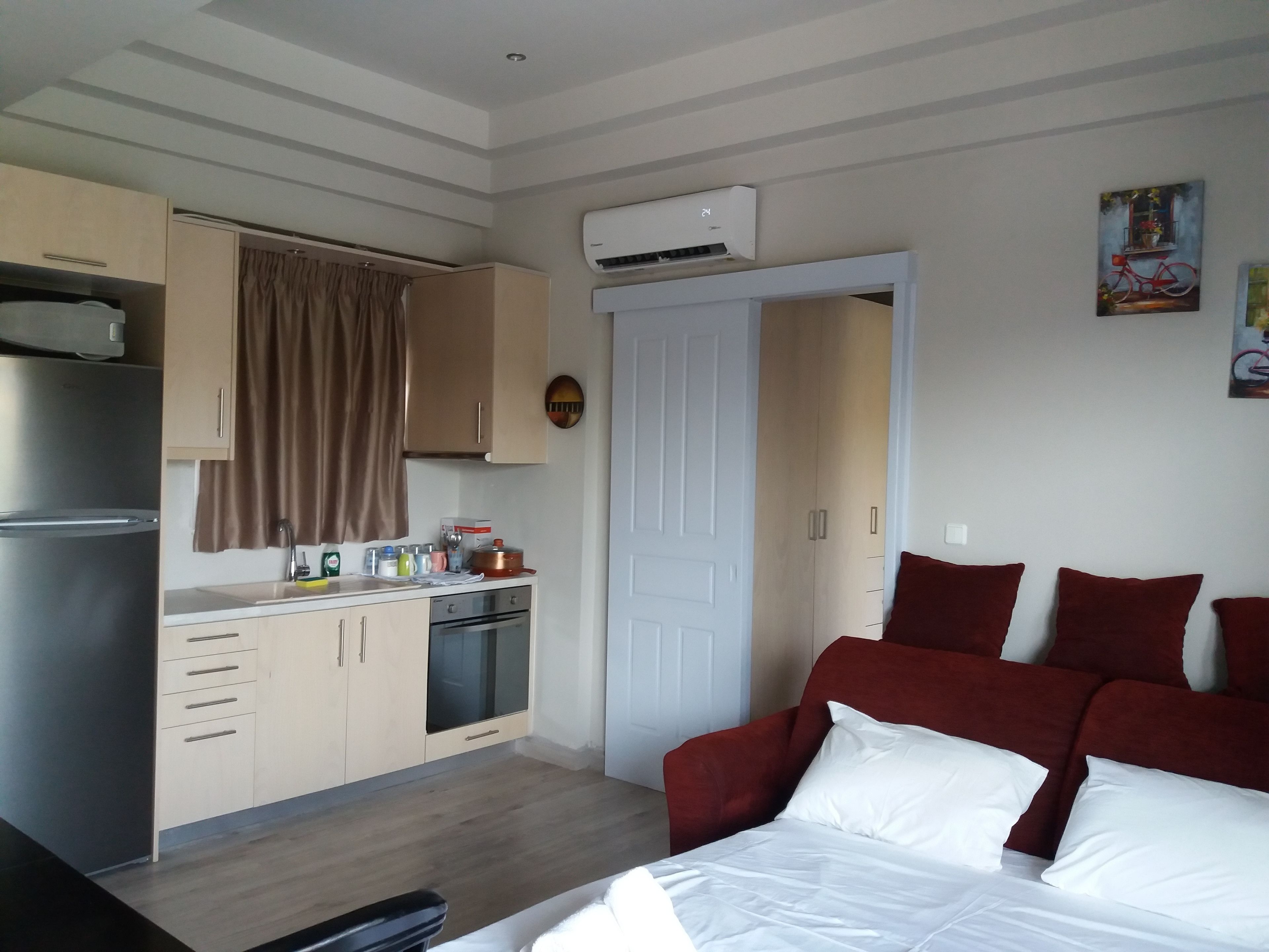A compact hotel room with a kitchenette, a bed with red cushions, and a painting on the wall.