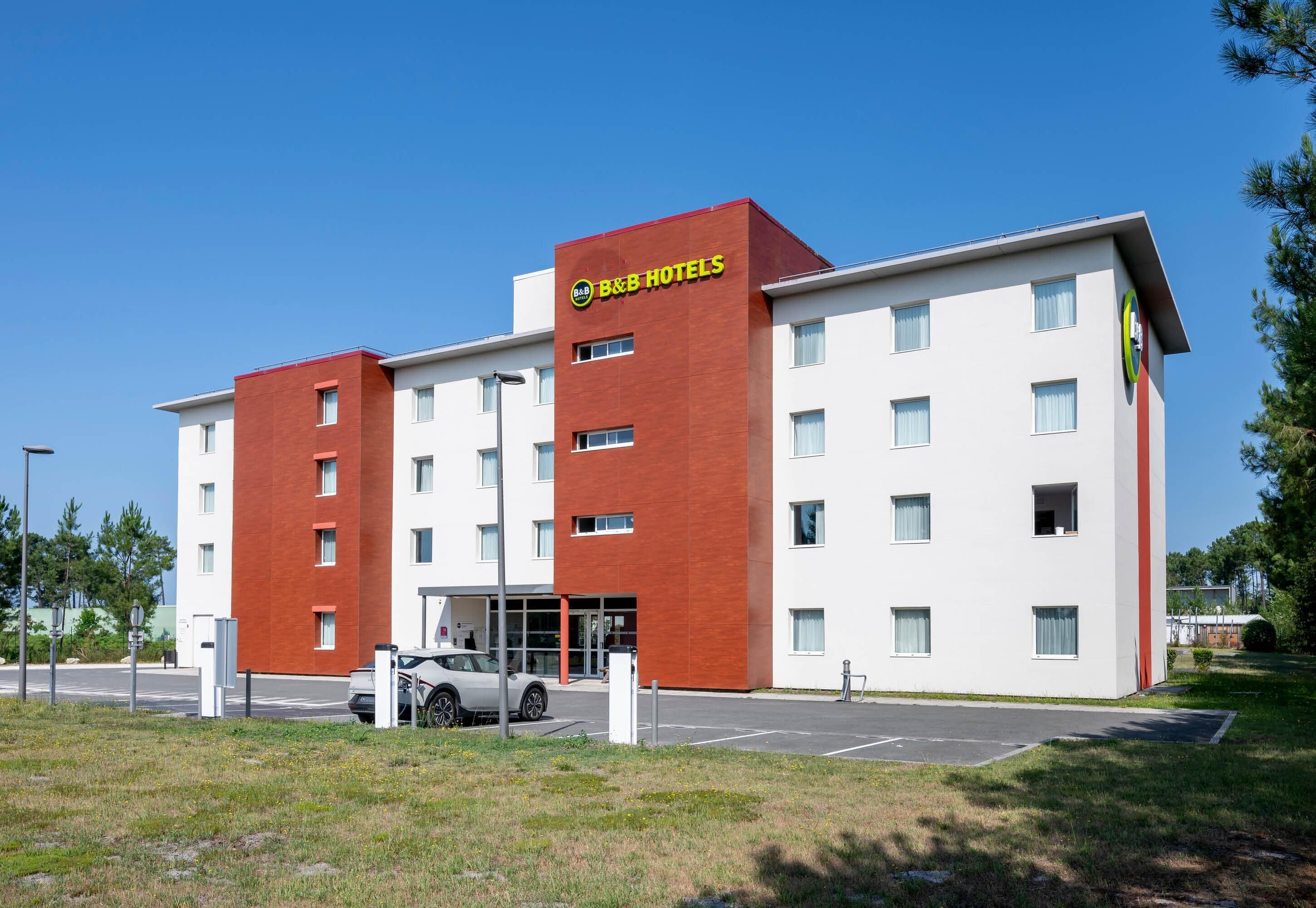 Un hotel tipo B&B con fachada roja y blanca, un estacionamiento y un cielo azul despejado.
