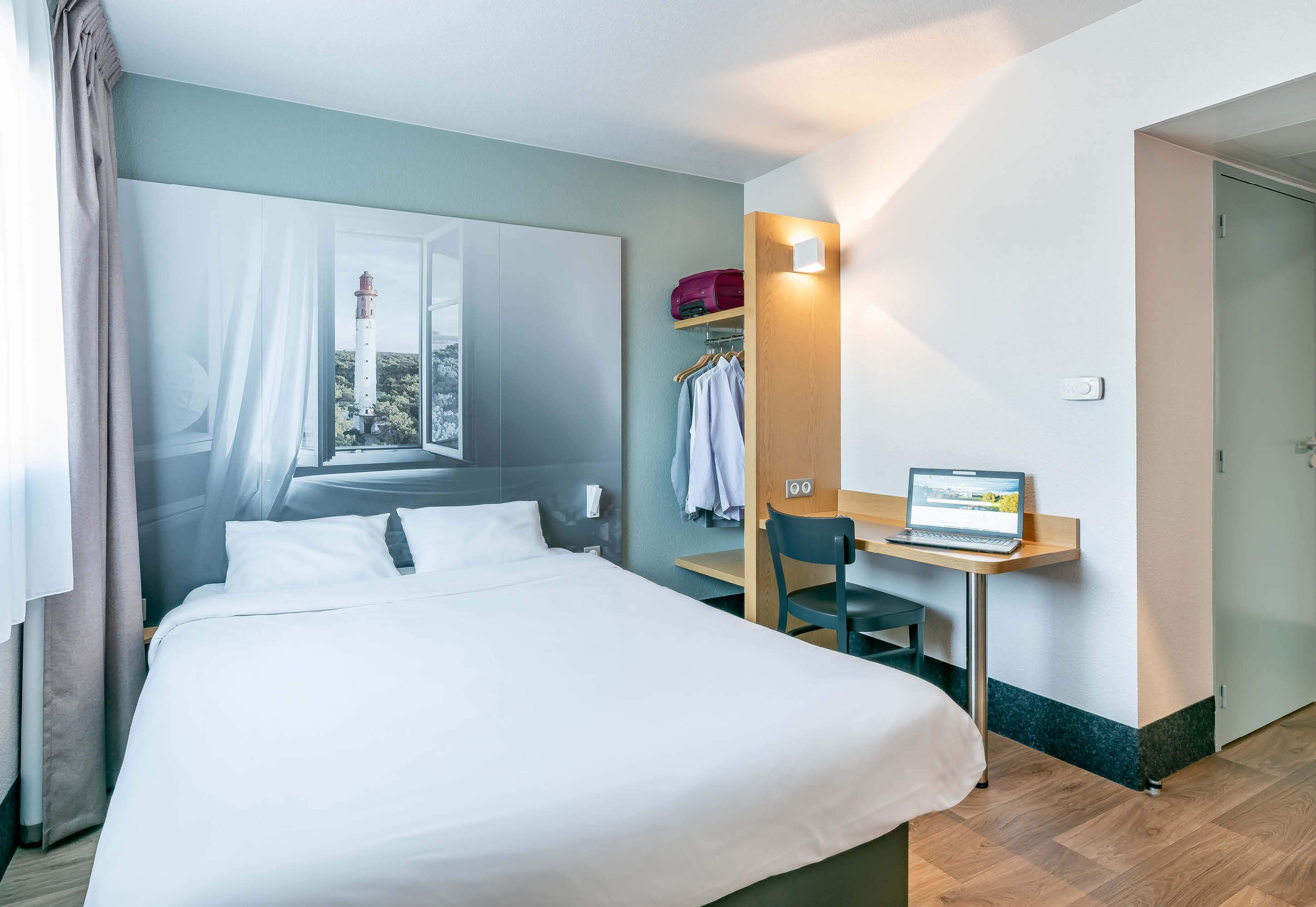 A hotel room with a bed, a desk with a laptop, a coat rack, and a view of a lighthouse through the window.