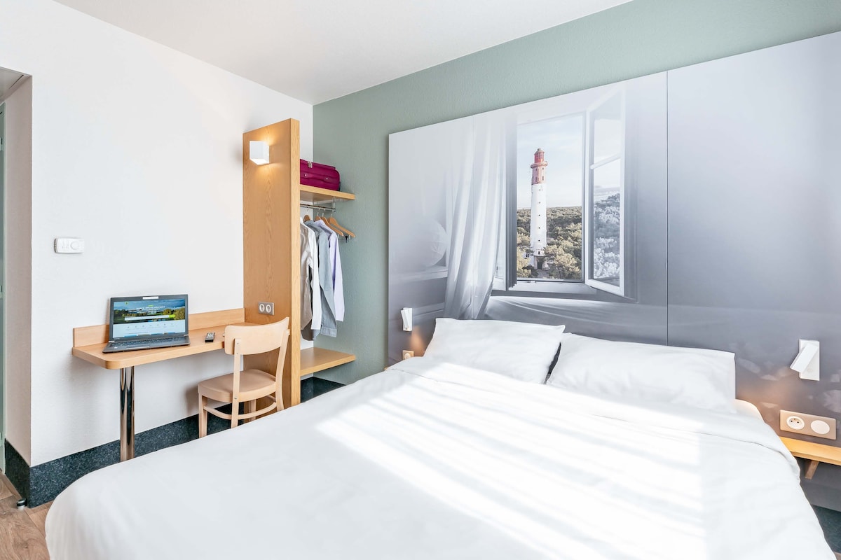 A hotel room with a bed, a desk with a laptop, a chair, a wardrobe, and a window with a view of a lighthouse.