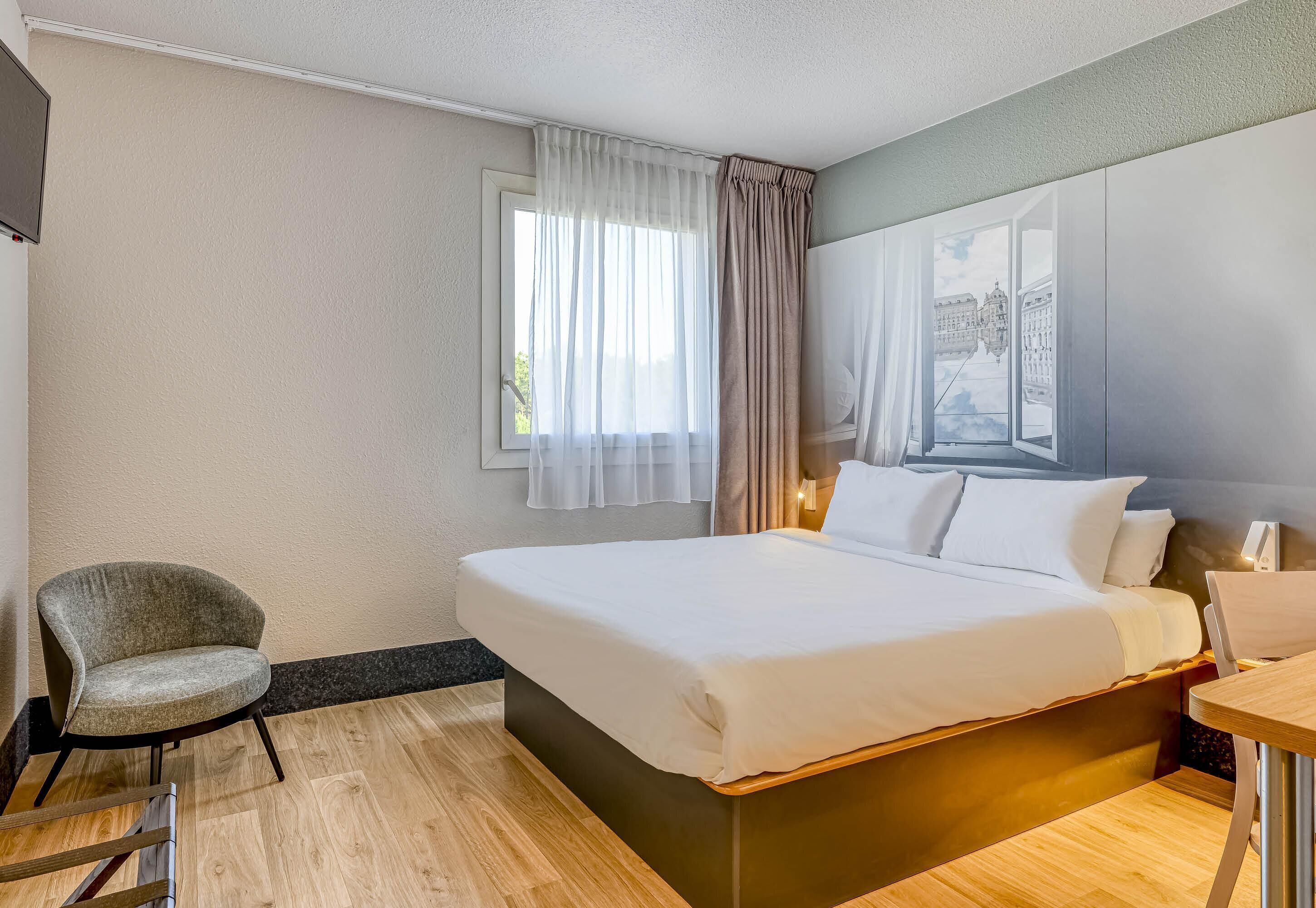 Una habitación de hotel con una cama, una silla, un escritorio y un ventanal con cortinas.