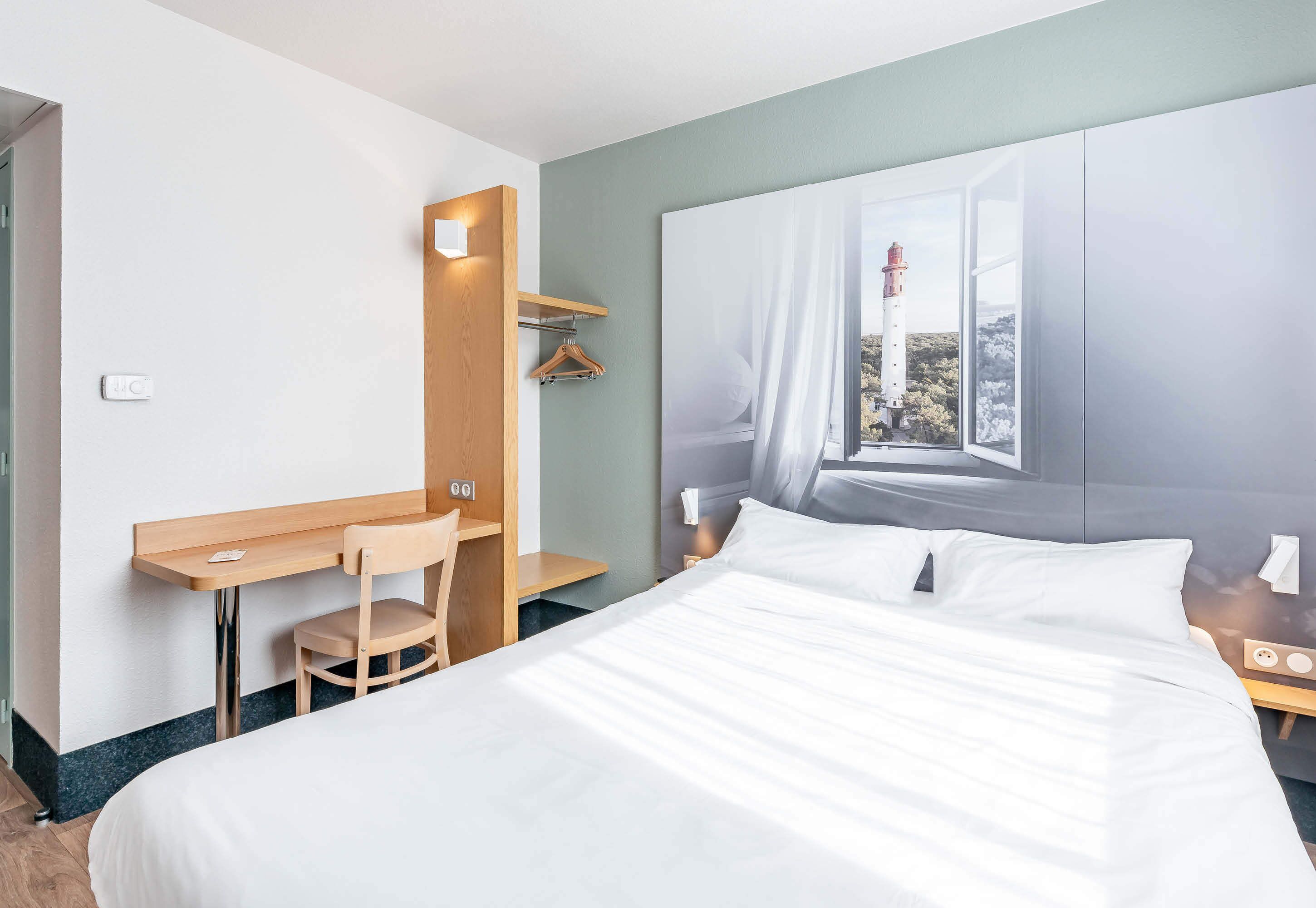 A hotel room with a bed, a desk, a chair, a window with a view of a lighthouse, and a nightstand.