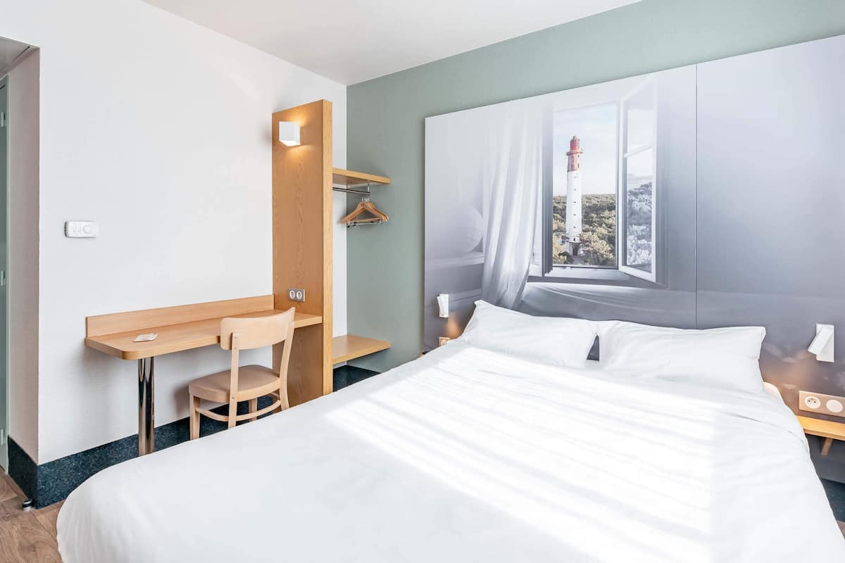 A hotel room with a bed, a desk, a chair, a window with a view of a lighthouse, and a nightstand.