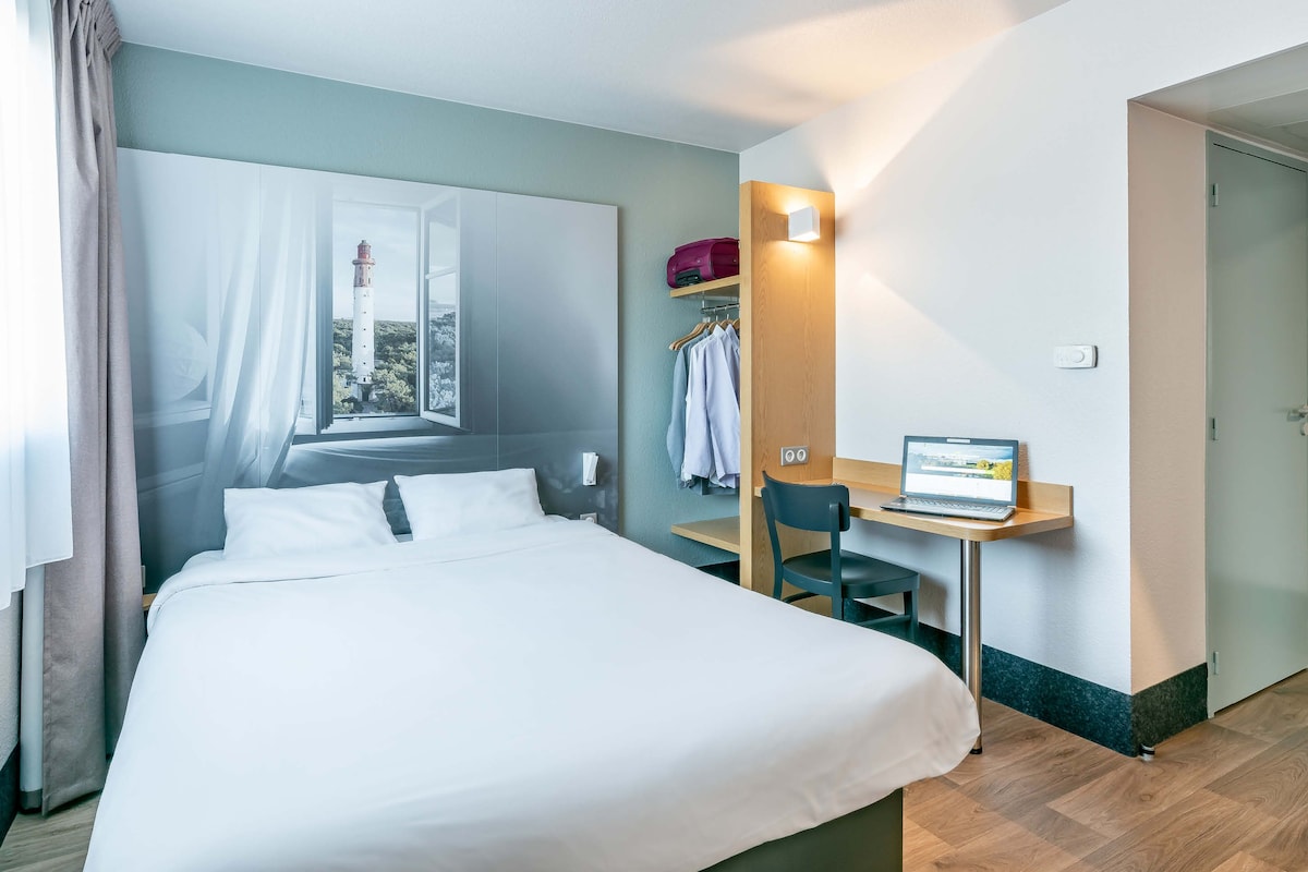 A hotel room with a bed, a desk with a laptop, a coat rack, and a view of a lighthouse through the window.