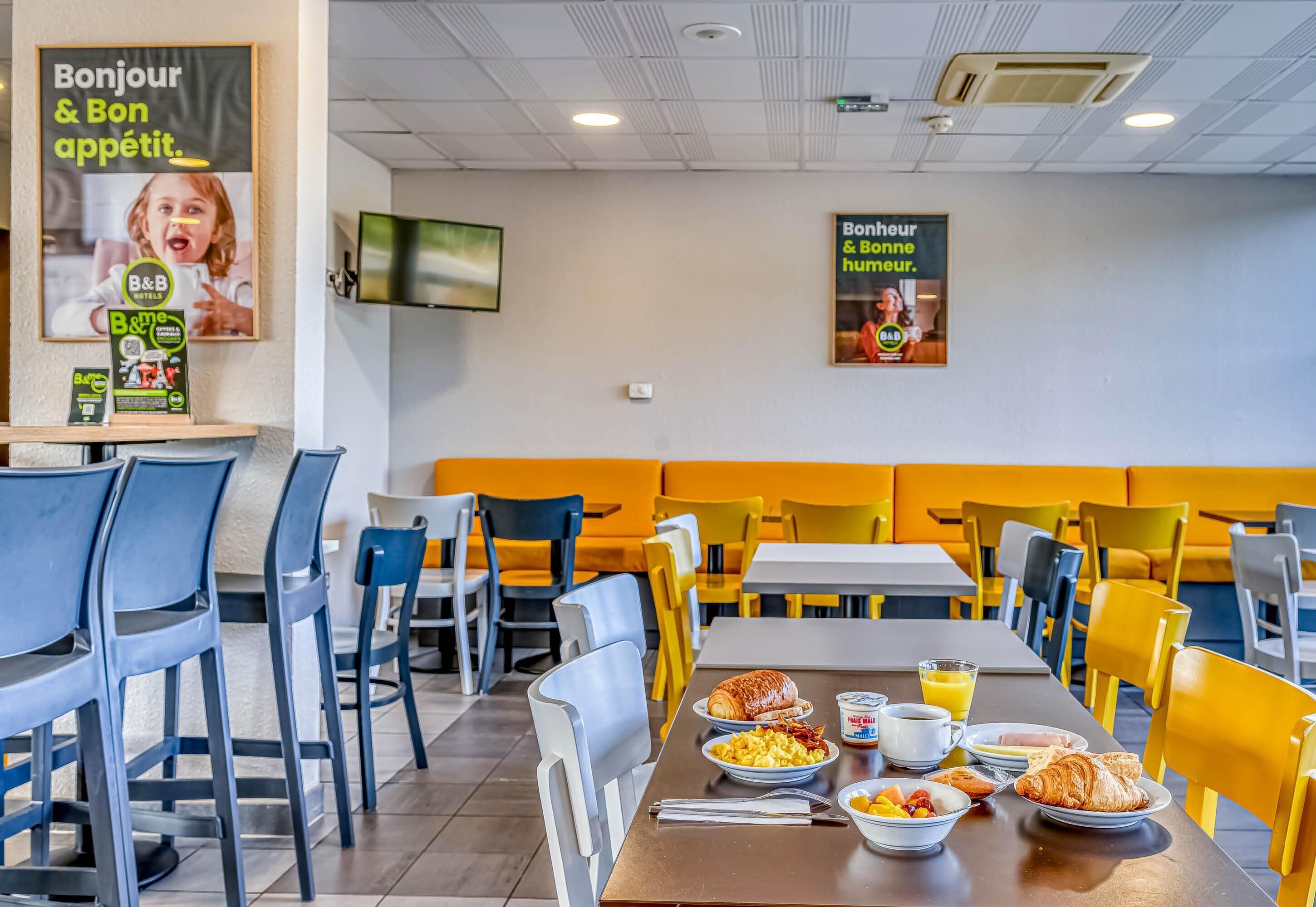 A restaurant with yellow and blue chairs, tables, and a breakfast spread including croissants, fruit, and beverages.