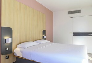 Siististi pedattu sänky hotellihuoneessa, jossa on puinen pääty, yöpöytävalaisimet ja valkoinen ovi.