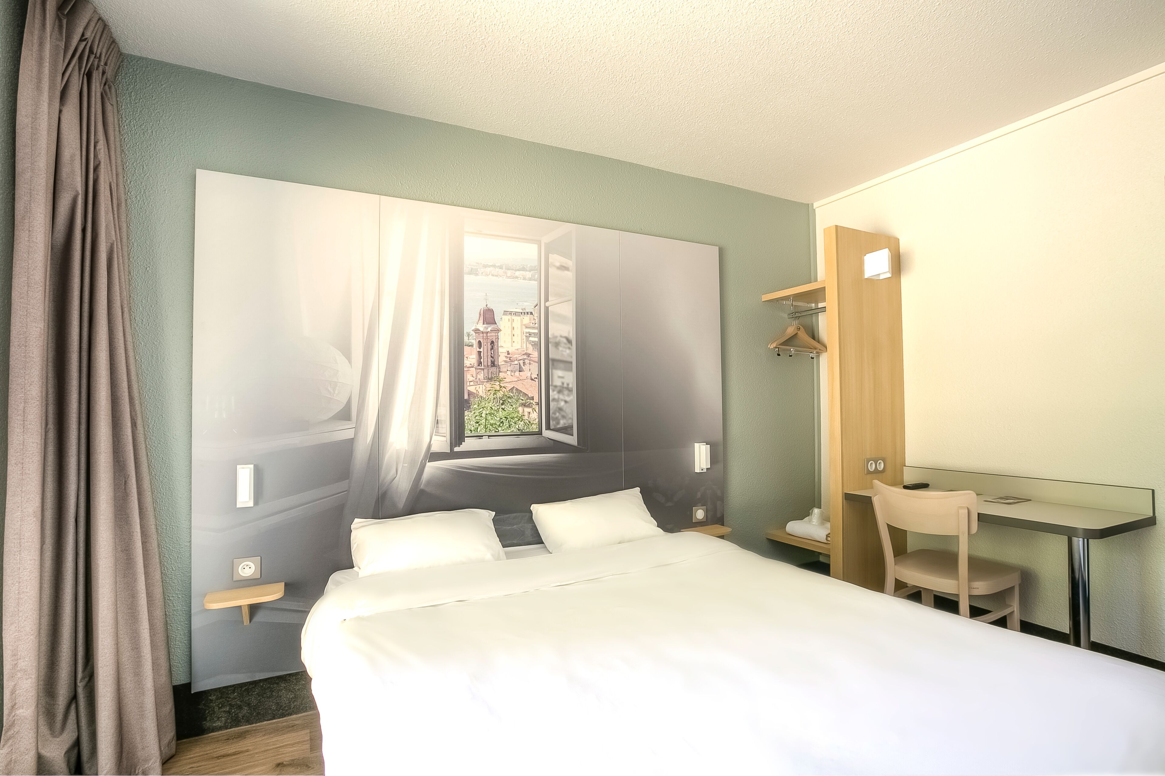 A hotel room with a large bed, a desk with a chair, a mirror, and a view of a cityscape through an open window.