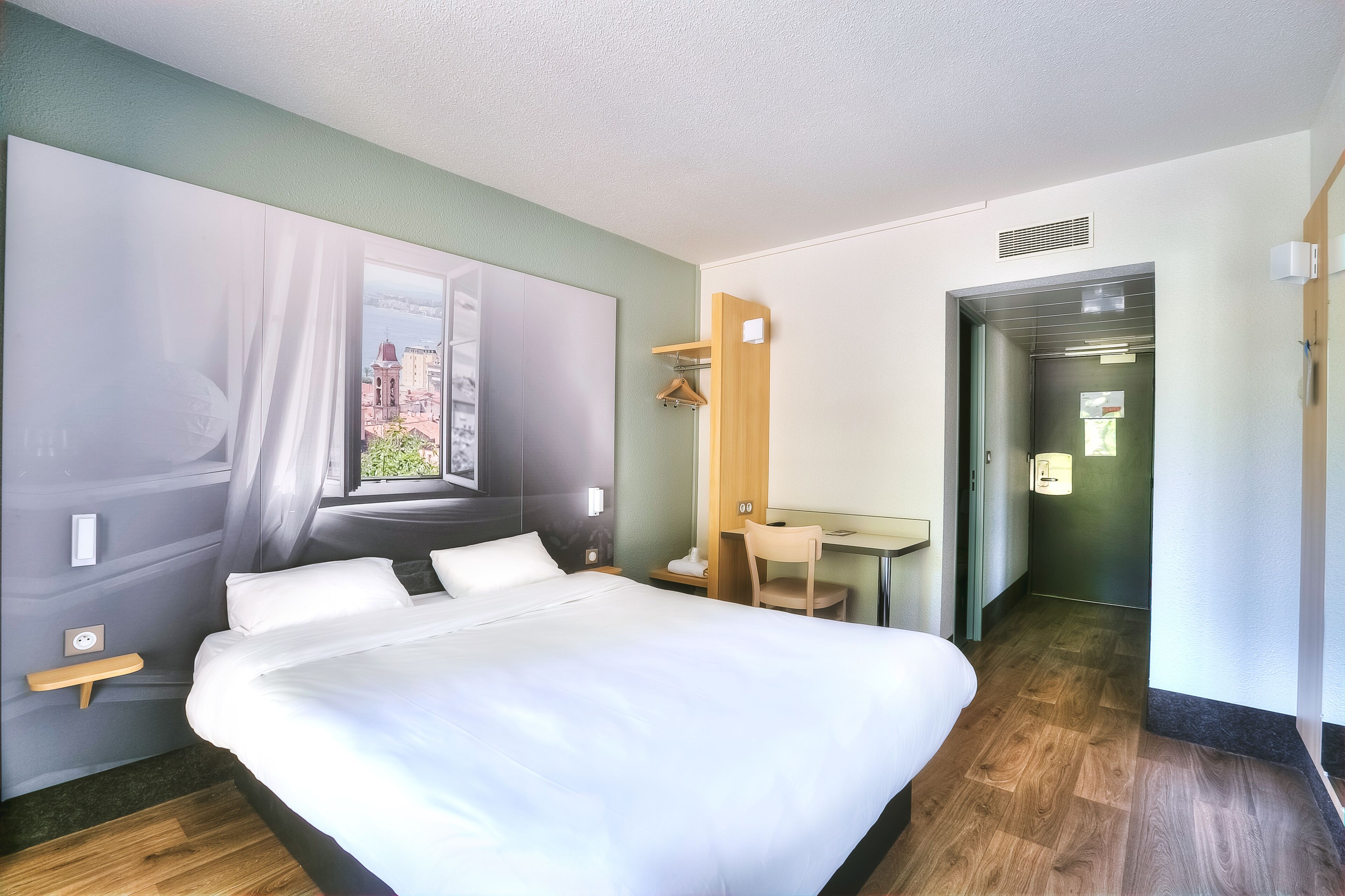 A modern hotel room with a large bed, a desk, and a view of a cityscape through sheer curtains.