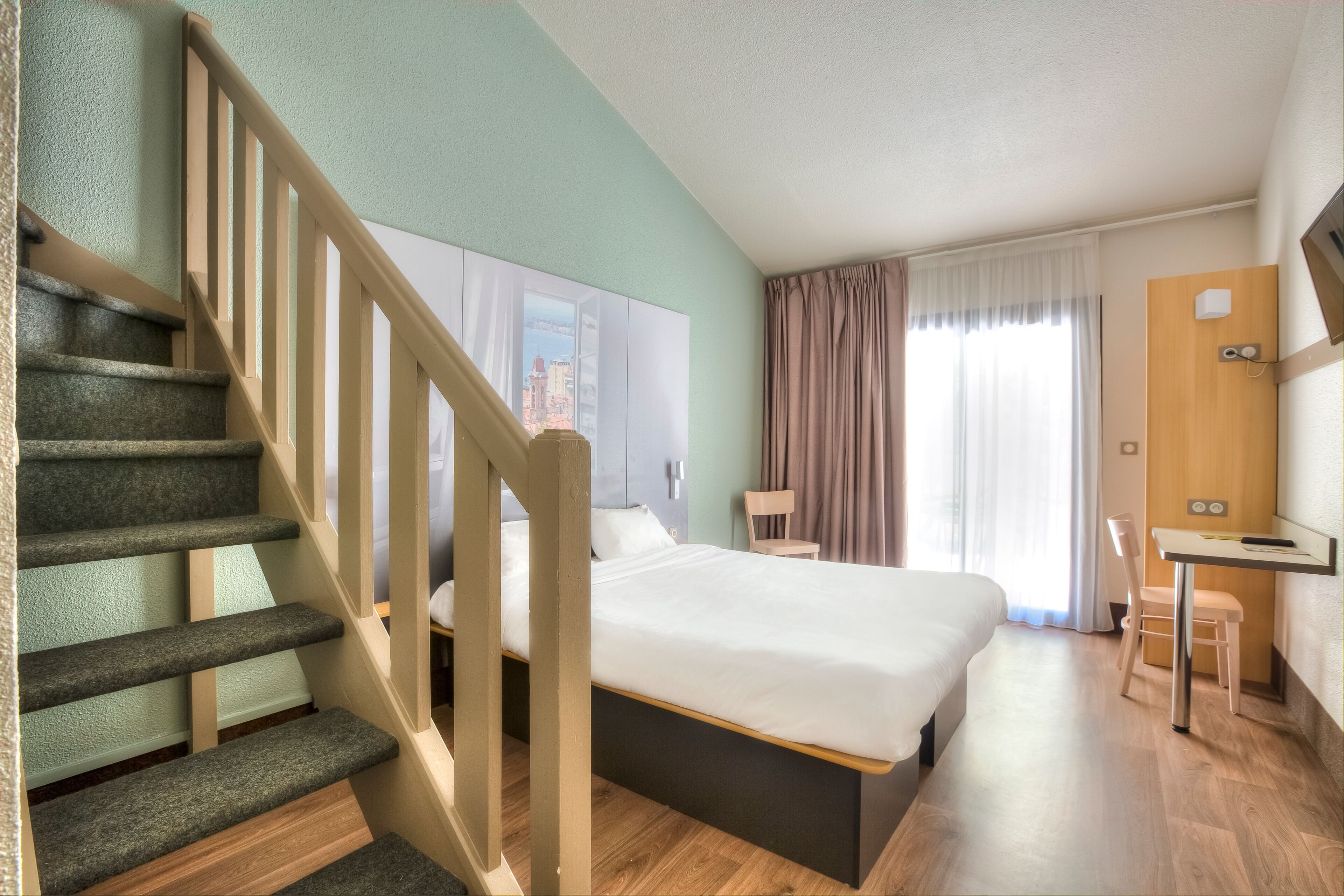 A hotel room with a bed, a desk, a chair, a staircase, and a view of a cityscape through a window.