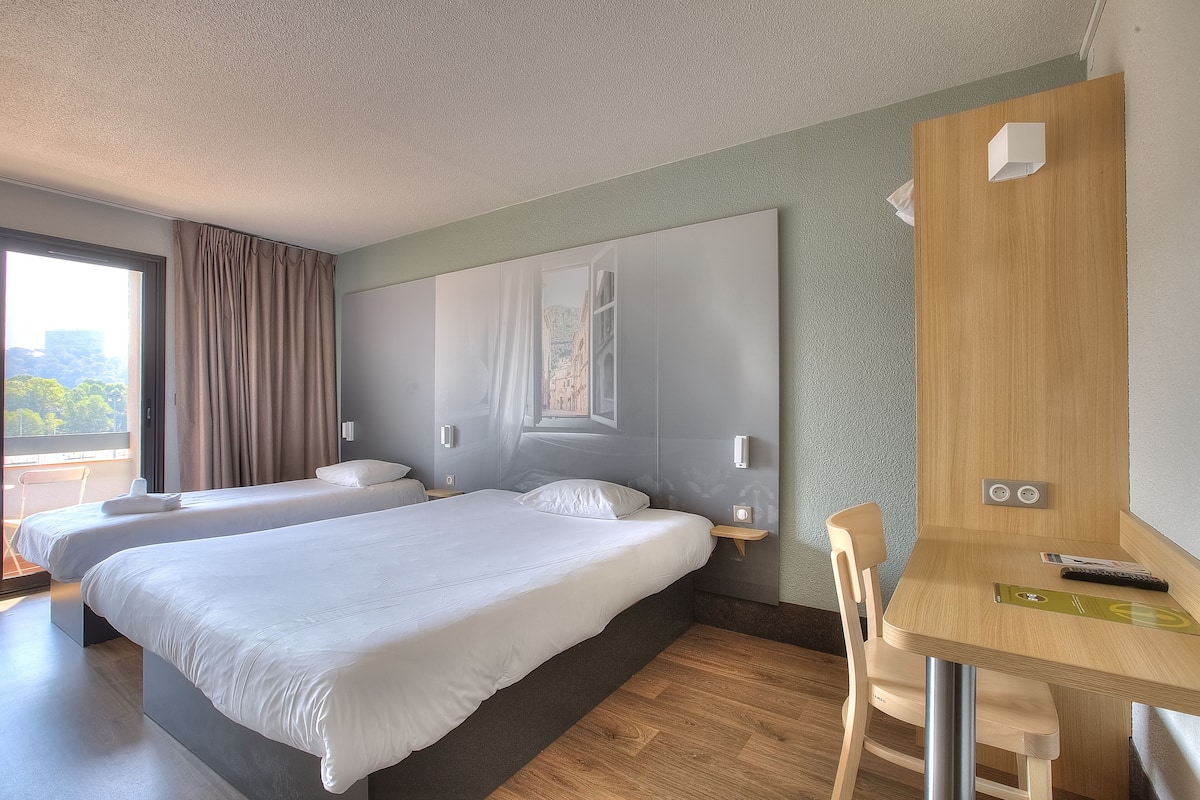 Habitación de hotel con dos camas, un escritorio de madera y vistas a un paisaje urbano a través de una ventana.
