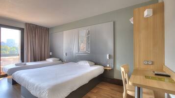 A hotel room with two beds, a wooden desk, and a view of a cityscape through a window.