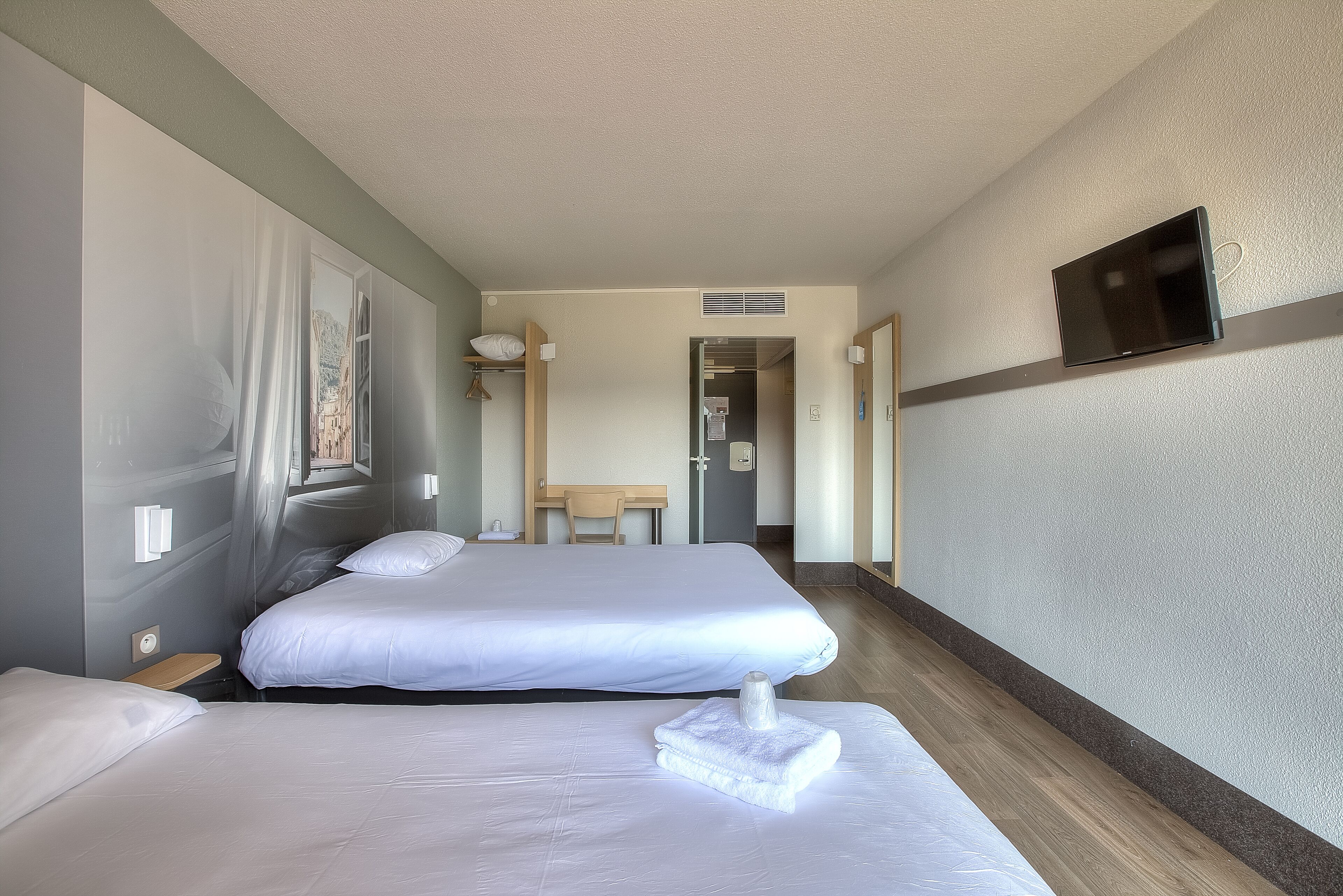 Habitació d'hotel amb dos llits, un televisor fixat a la paret i vista d'un edifici pel finestral.