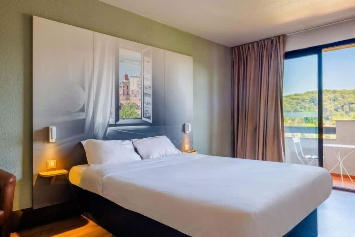 Habitación de hotel con cama, mesita de noche, ventana con cortinas y vistas a un balcón.