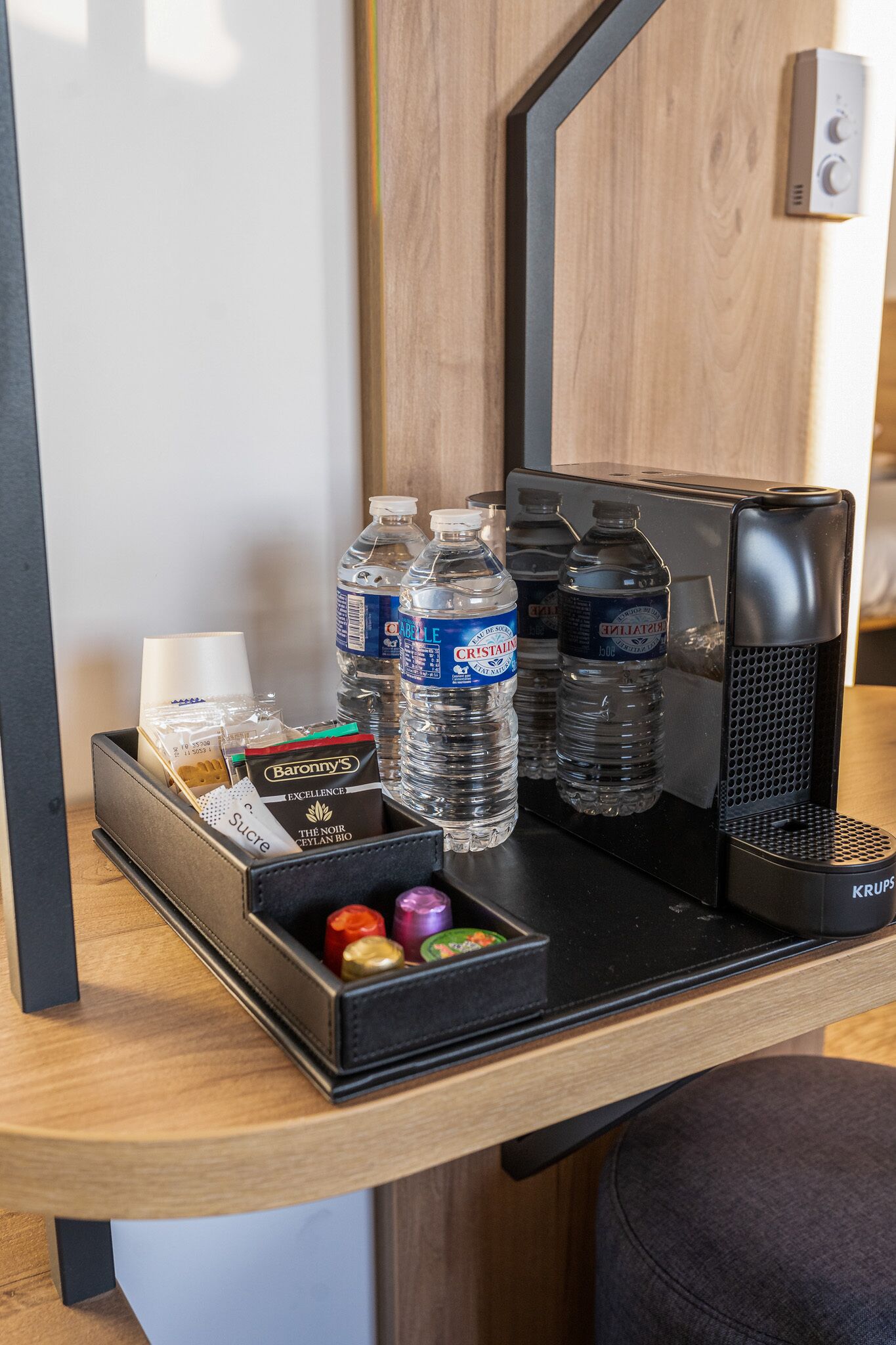 Sur le bureau de la chambre d’hôtel, on trouve une machine à café, deux bouteilles d’eau, un paquet de sucre et une boîte de chocolats.