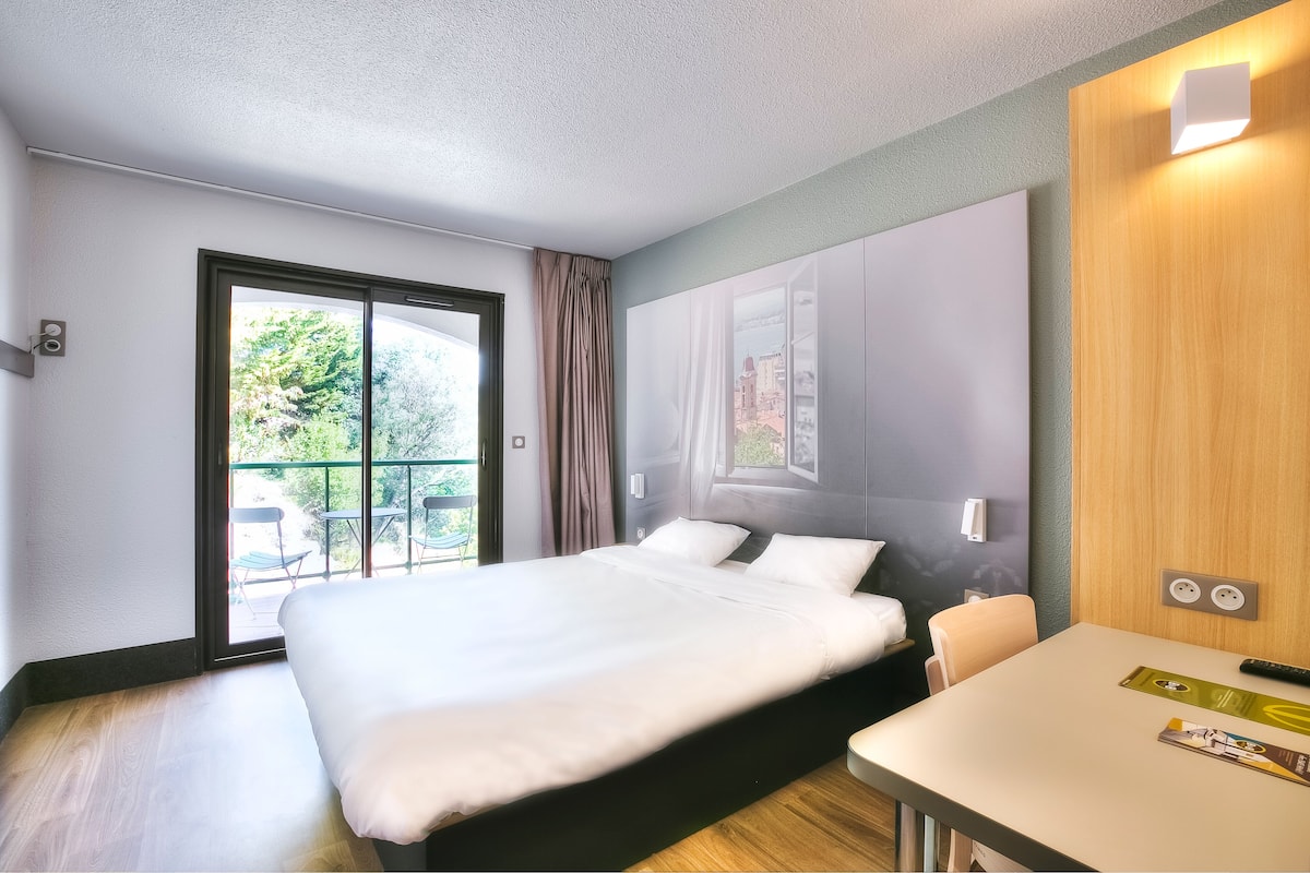 Una habitación de hotel moderna con una cama grande, una mesita pequeña y vistas al exterior a través de una puerta corredera.