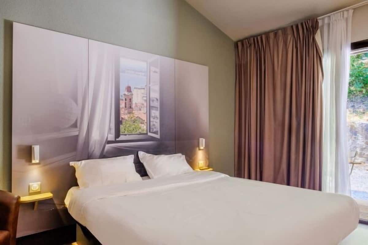 Habitación de hotel con una cama grande, dos lámparas de noche, cabecera con una obra de arte de un paisaje urbano y una ventana con cortinas.