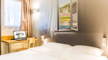 Een hotelkamer met een bed, een bureau met een laptop, een raam met uitzicht op een rivier en gebouwen, en gordijnen.