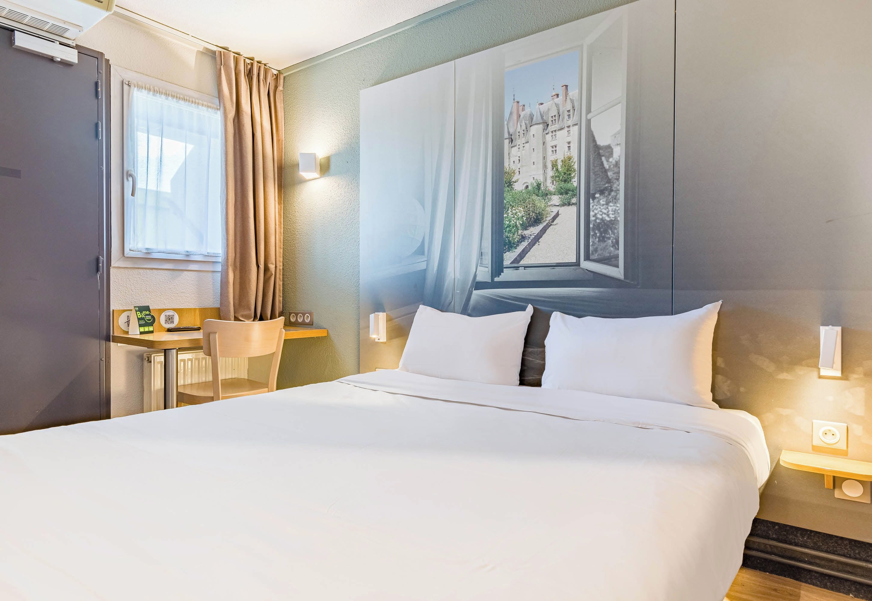 A hotel room with a large bed, a desk with a chair, a window with curtains, and a wall-mounted light.