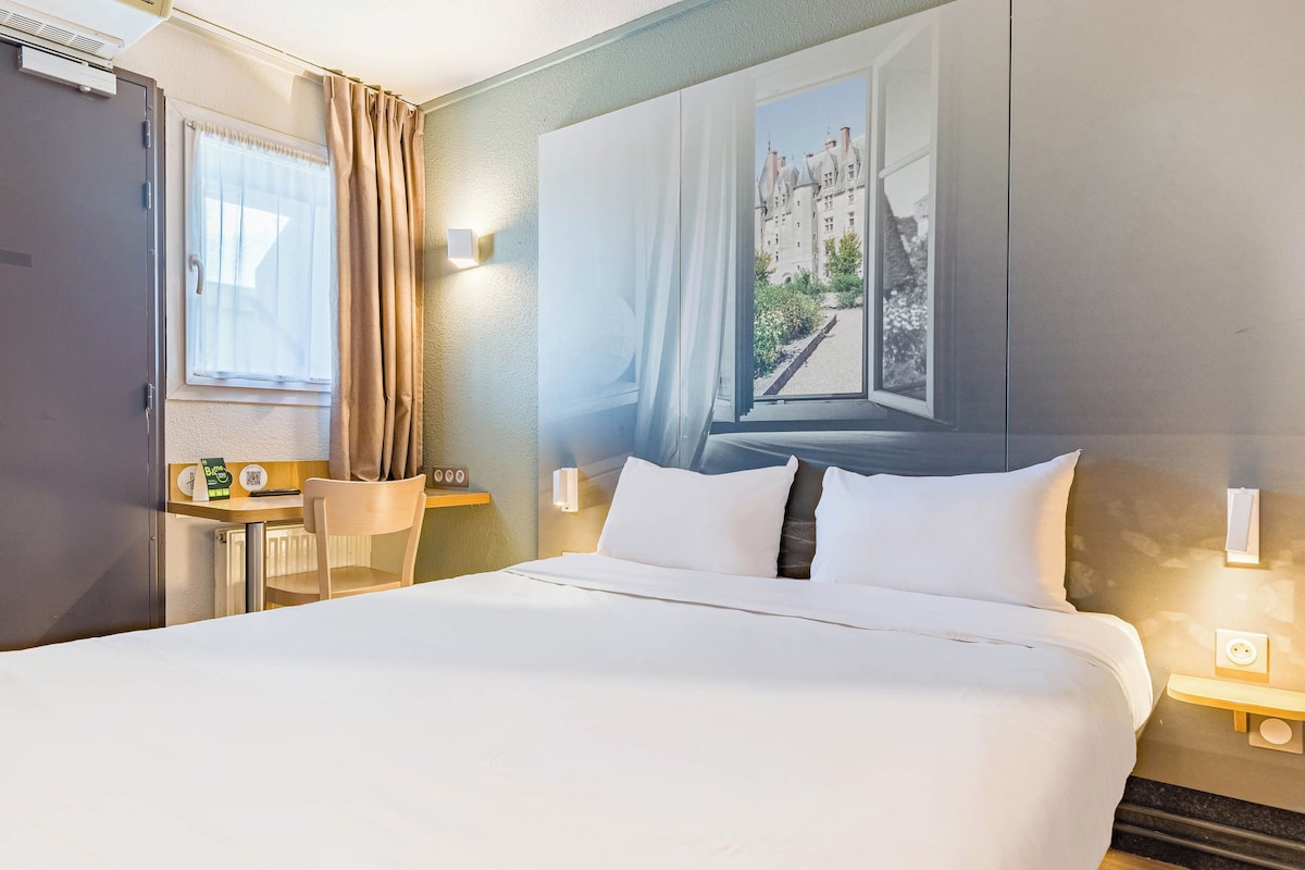 Une chambre d’hôtel avec un grand lit, un bureau avec une chaise, une fenêtre avec des rideaux et un lampadaire fixé au mur.