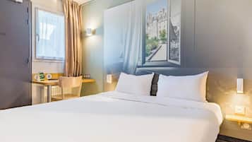 A hotel room with a large bed, a desk with a chair, a window with curtains, and a wall-mounted light.