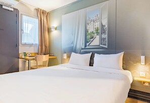 A hotel room with a large bed, a desk with a chair, a window with curtains, and a wall-mounted light. - B&B Hotel Tours Sud Joué-lès-tours (Joue-les-Tours)