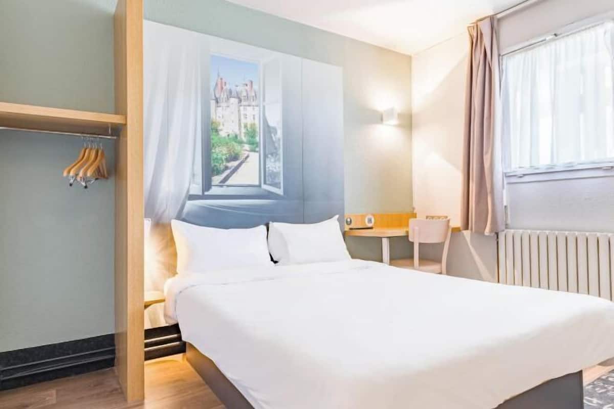 Ein Hotelzimmer mit Bett, einem Fenster mit Blick auf Gebäude, einem kleinen Tisch mit Stuhl und einem Kleiderständer mit Kleiderbügeln.
