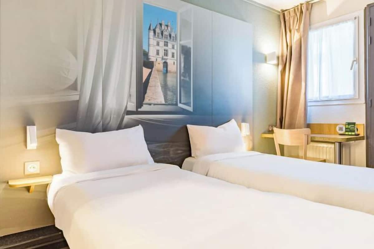 Ein Hotelzimmer mit zwei Betten, einem Fenster mit Vorhängen, einem Nachttisch und einer Wandlampe.