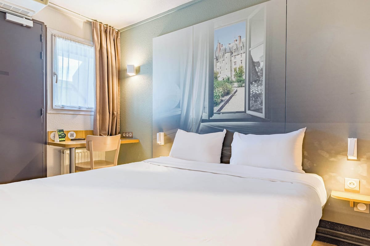 Ein Hotelzimmer mit einem großen Bett, einem Schreibtisch mit Stuhl, einem Fenster mit Vorhängen und einer Wandleuchte.