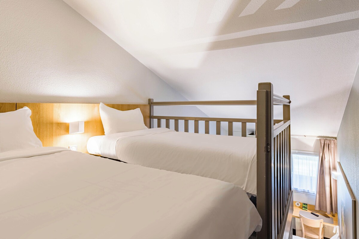 Ein Hotelzimmer mit zwei Betten, Holz-Kopfteilen, einem kleinen Tisch und einem Fenster mit Vorhängen.
