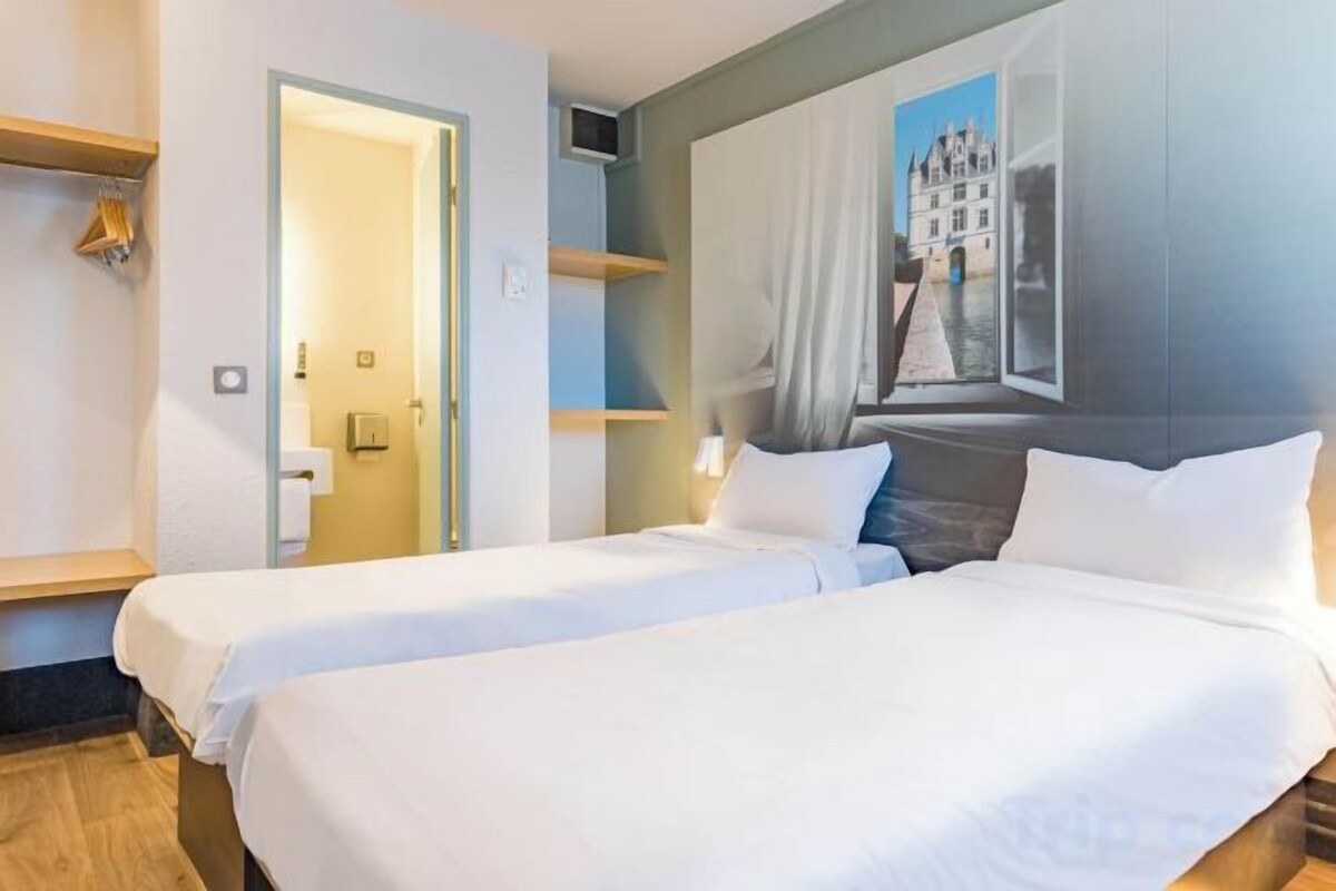A hotel room with two beds, a painting of a cityscape, and a door leading to a bathroom.