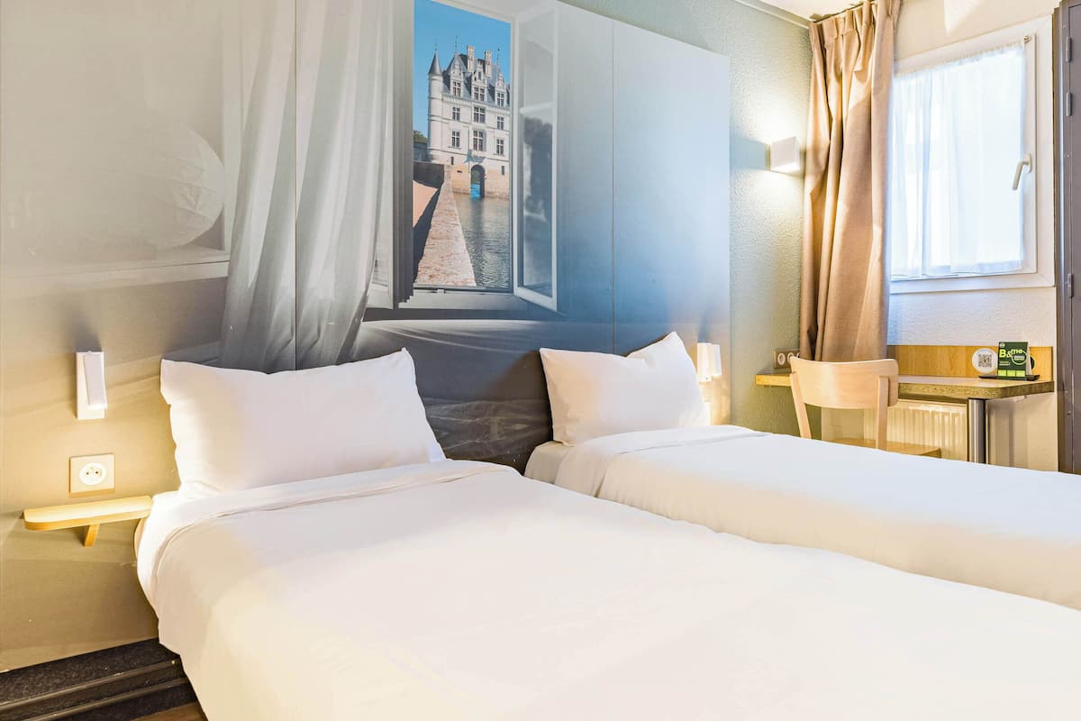 Ein Hotelzimmer mit zwei Betten, einem großen Fenster mit Vorhängen, einer Wanduhr und einem kleinen Tisch mit Lampe.