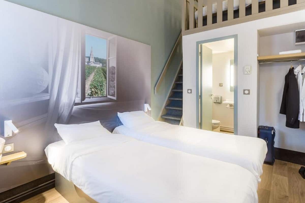 A hotel room with two beds, a window with a view, a staircase, and a coat rack.